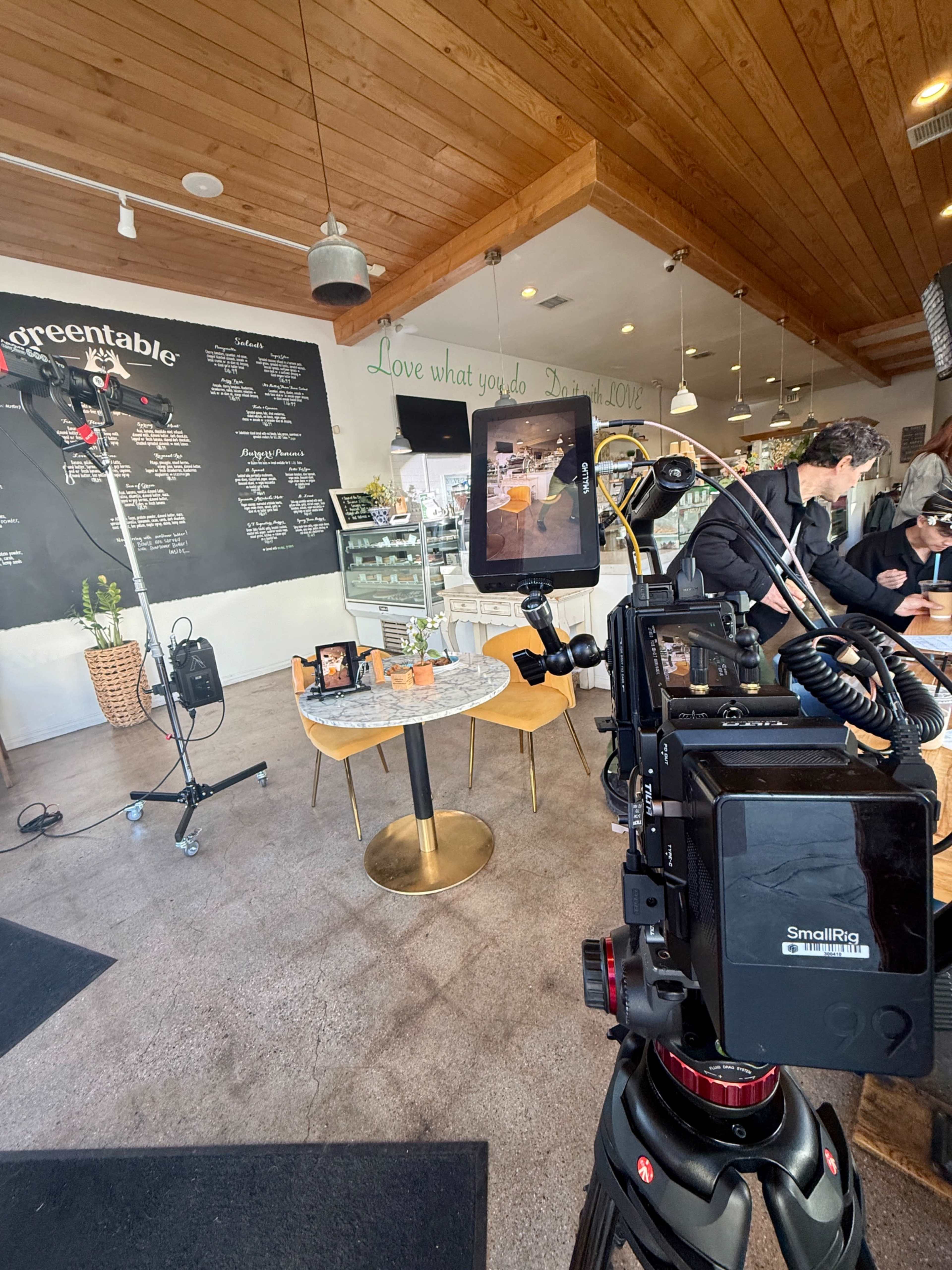 A camera is set up on a tripod facing a table in a café, with a light stand in the background and patrons at the counter.
