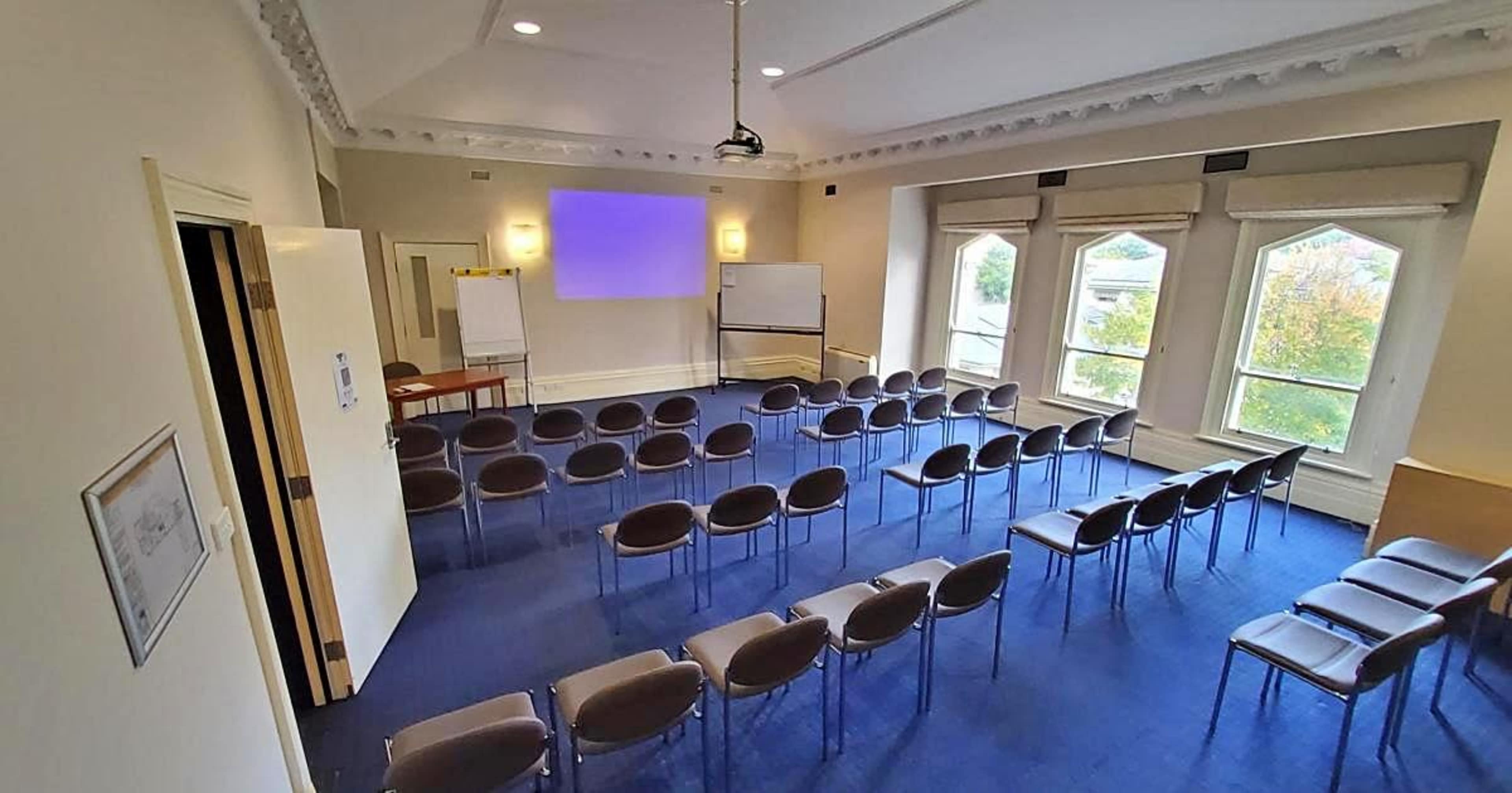 The image shows a conference room with rows of chairs facing a blank projection screen, flanked by windows and a table at the back.
