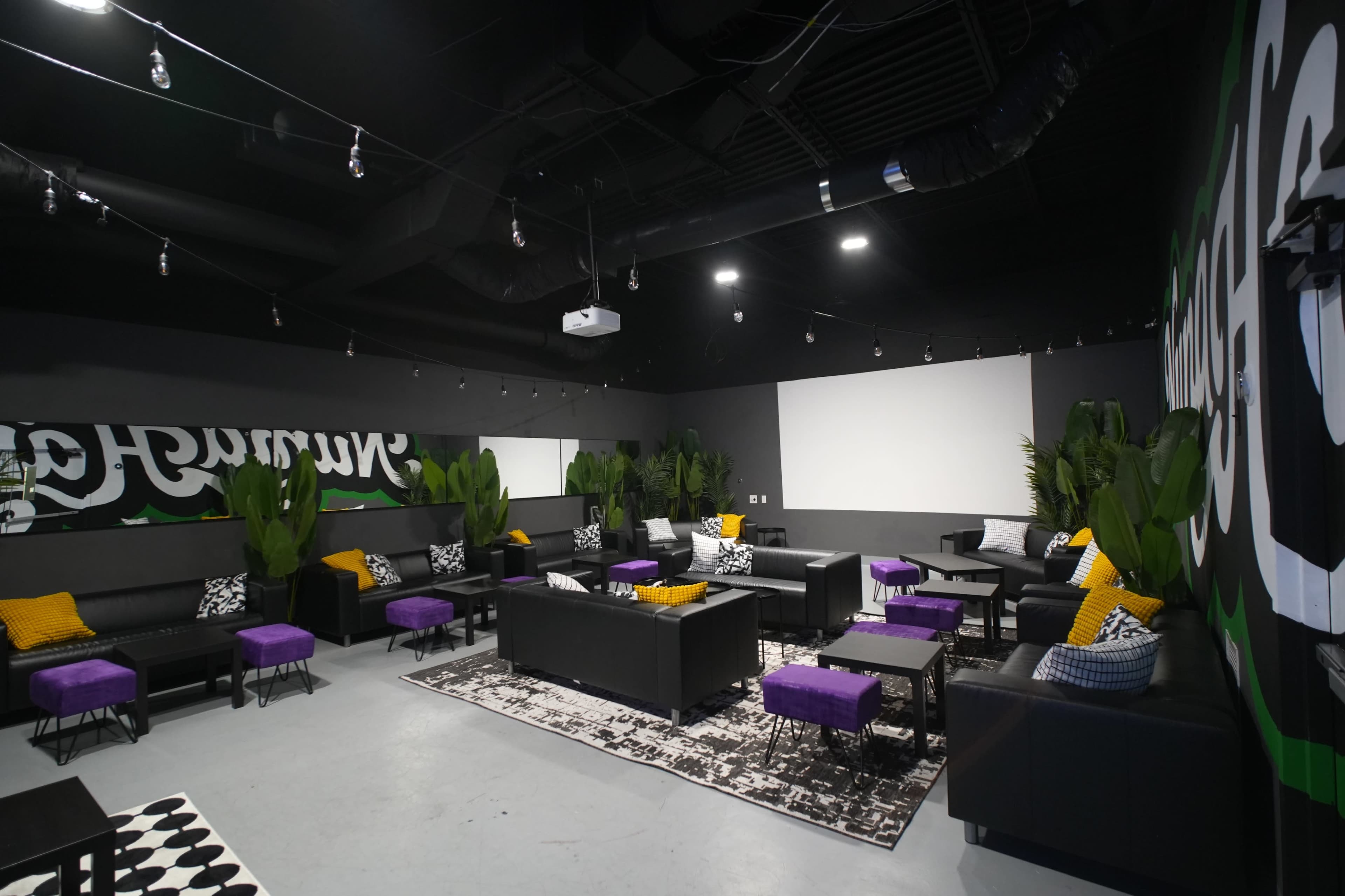 The image shows a modern lounge area featuring black seating, purple accent pillows, and large plants, with a mural on the wall and a projector screen.