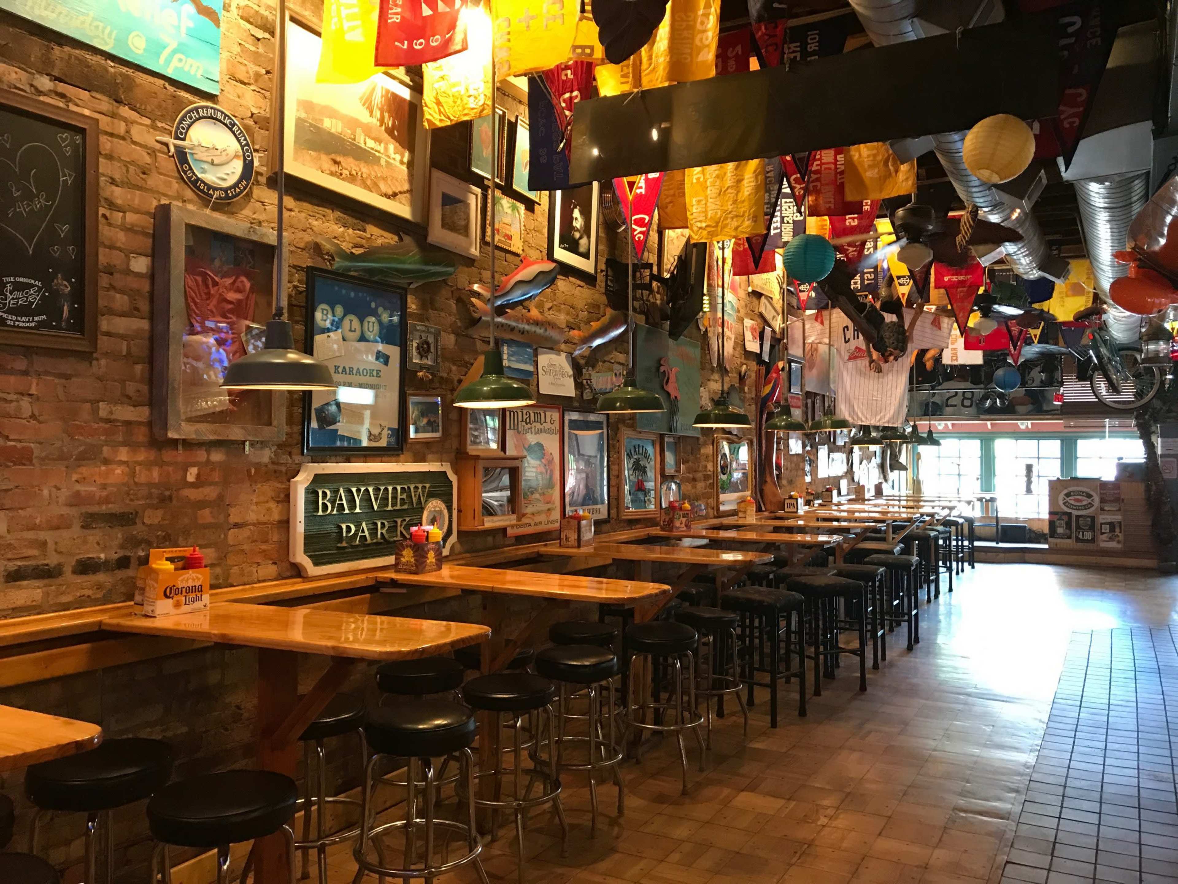 The image shows an interior of a bar or restaurant with wooden tables and stools, brick walls adorned with various decorations, including flags and photographs.