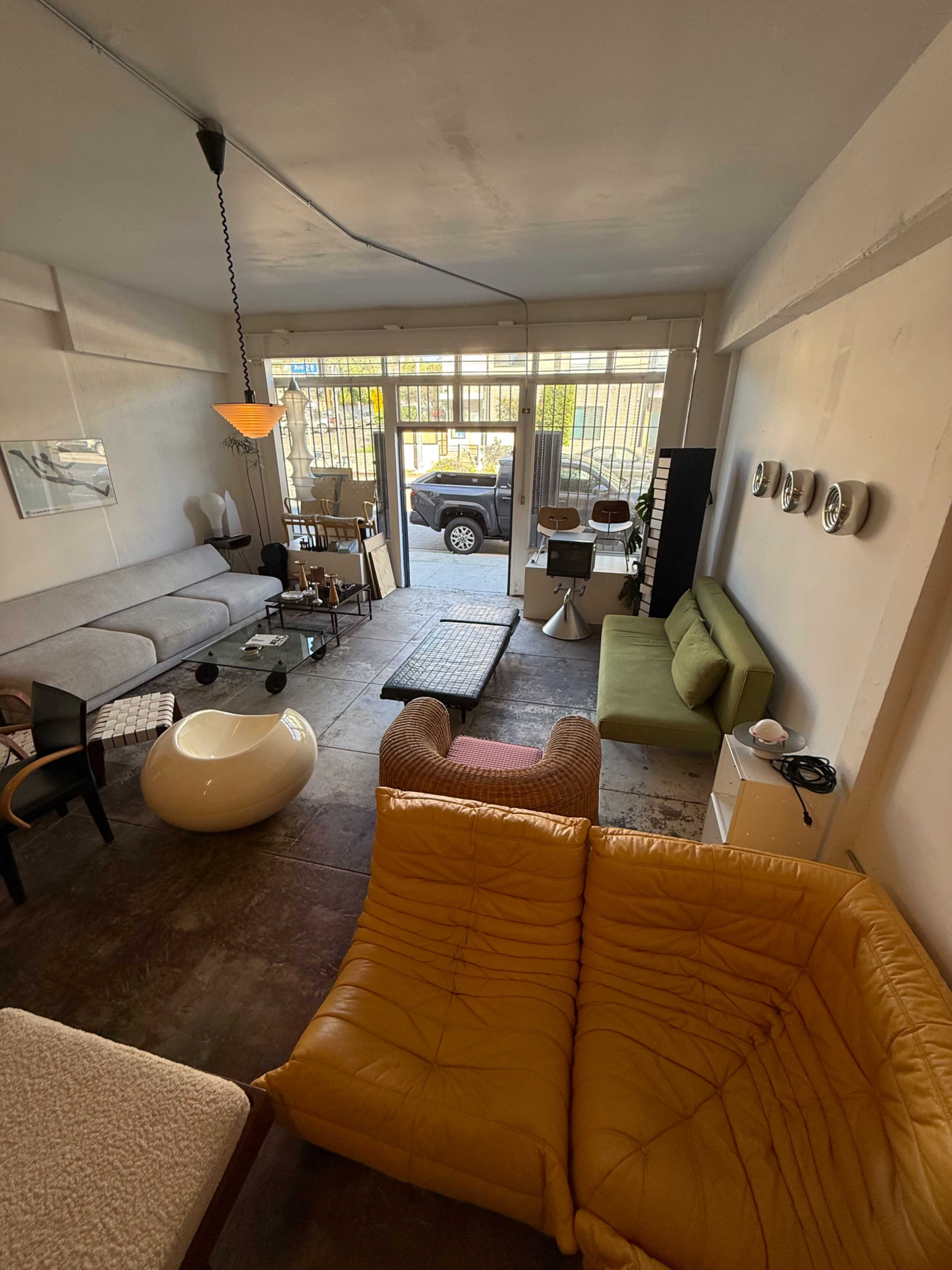 The image shows a modern interior space with various seating arrangements, a large window overlooking a street, and a mix of furniture including a yellow couch, a green sofa, and an overstuffed chair.