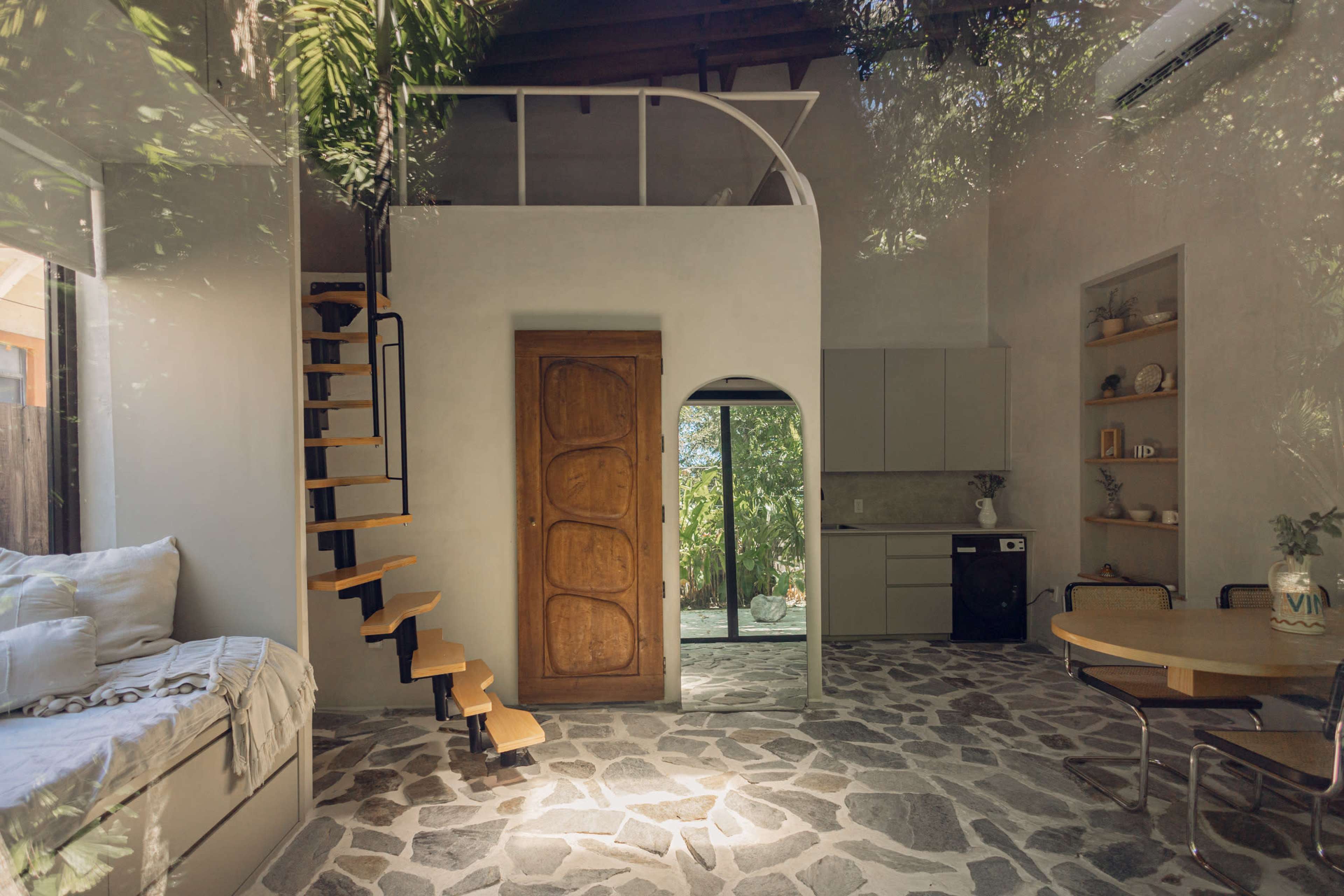 The image shows a modern, open-plan living space featuring a wooden staircase, a kitchenette, and large windows that lead to an outdoor area.