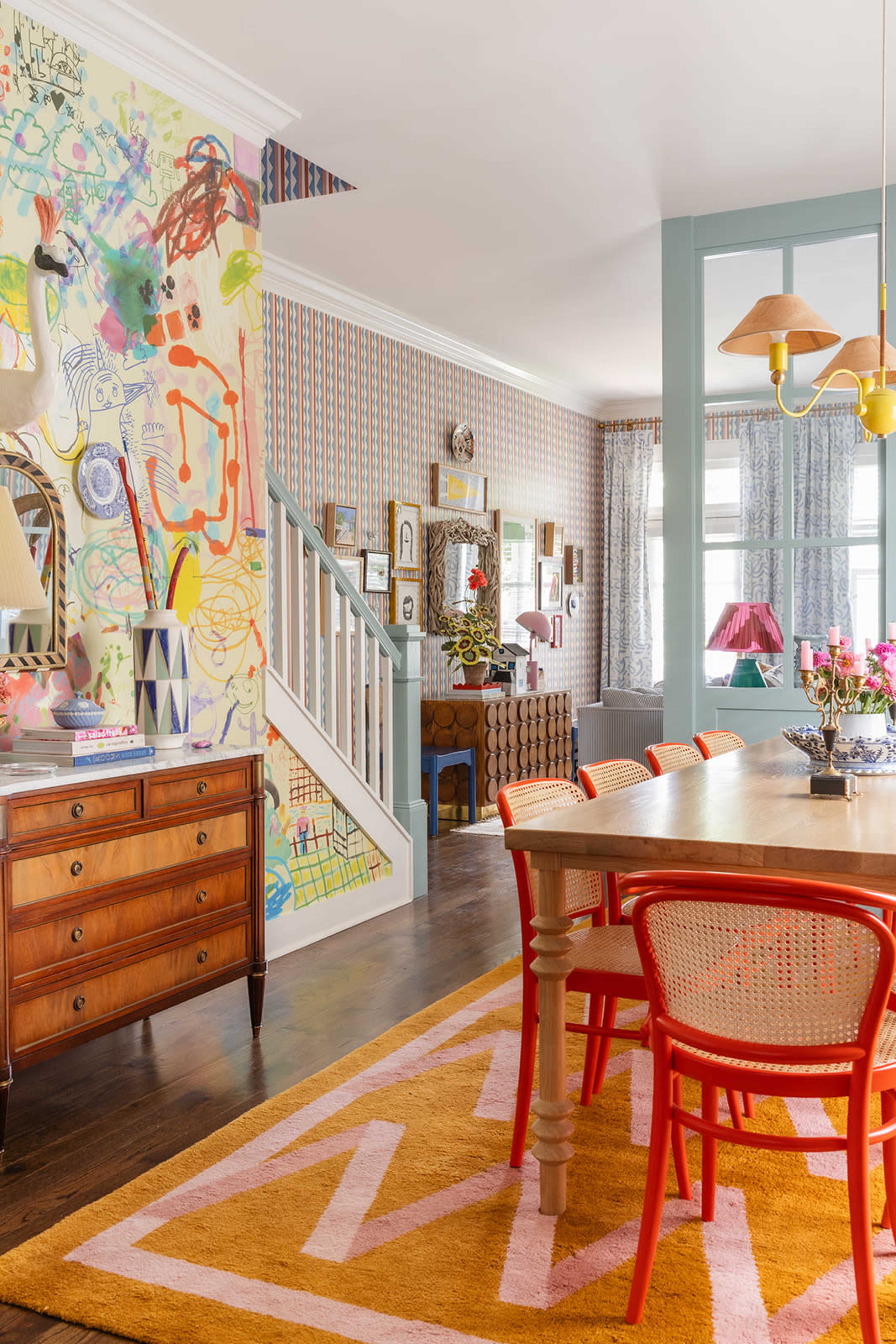 The image depicts a colorful dining area with a wooden table surrounded by red chairs, vibrant patterned wallpaper, and a staircase in the background.