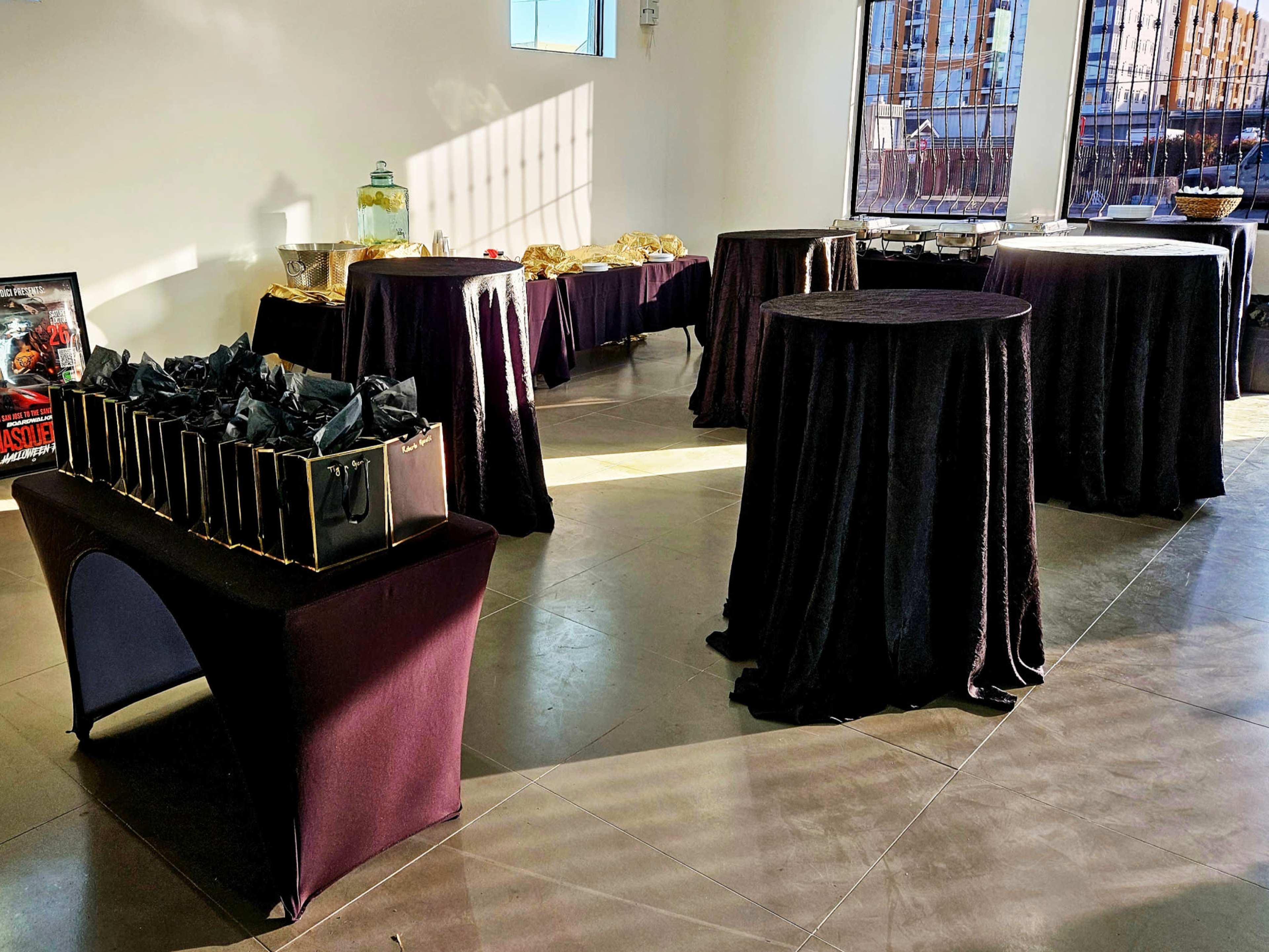 The image shows a spacious event room set with several round tables covered in dark cloth, featuring food displays and gift bags arranged on a table against the wall.