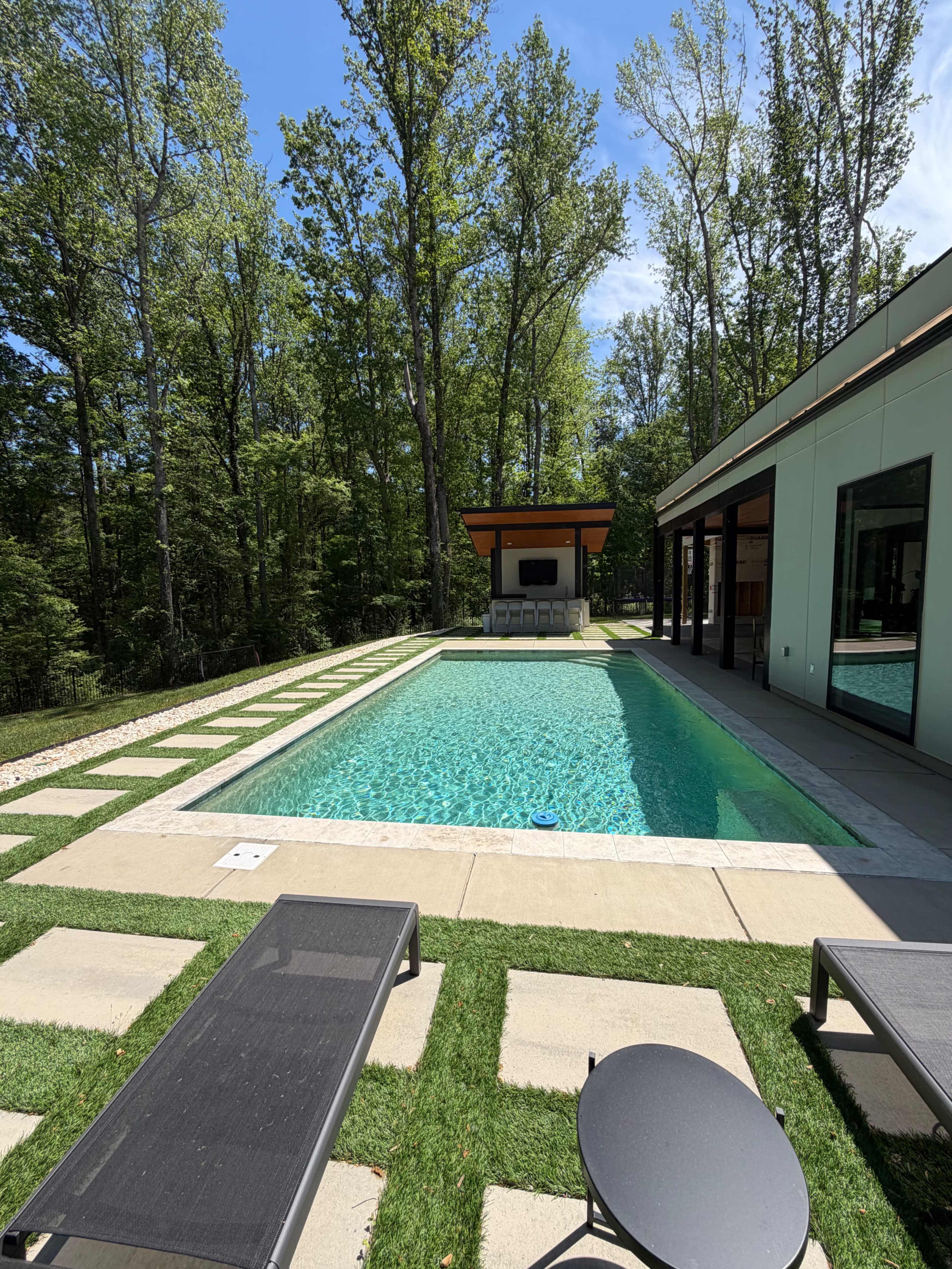 The image shows a modern swimming pool surrounded by concrete pavers and lush green grass, set against a backdrop of tall trees and a contemporary building with large windows.