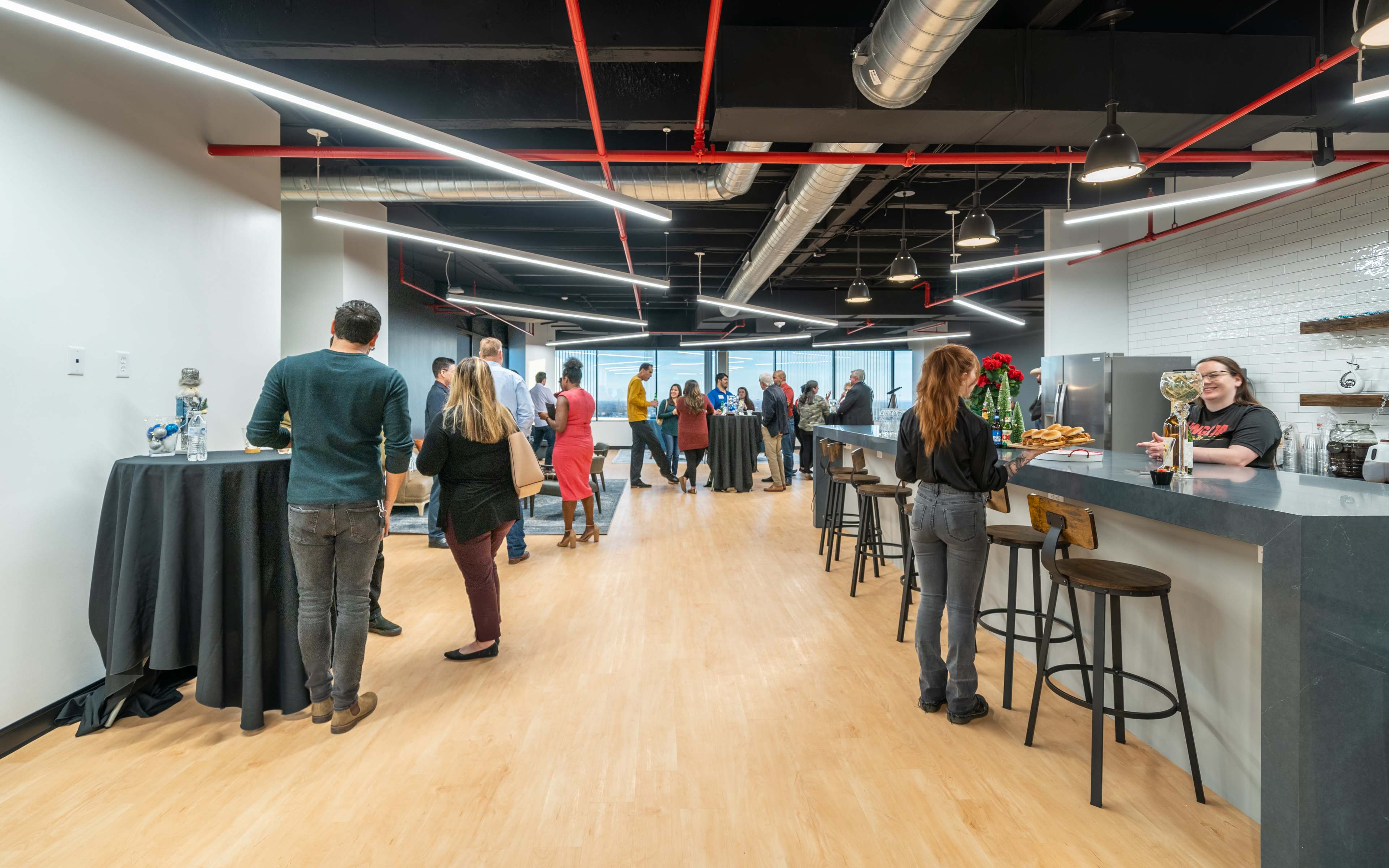 A group of people gathers in an office setting with tables and a bar, engaging in conversation near a large window.