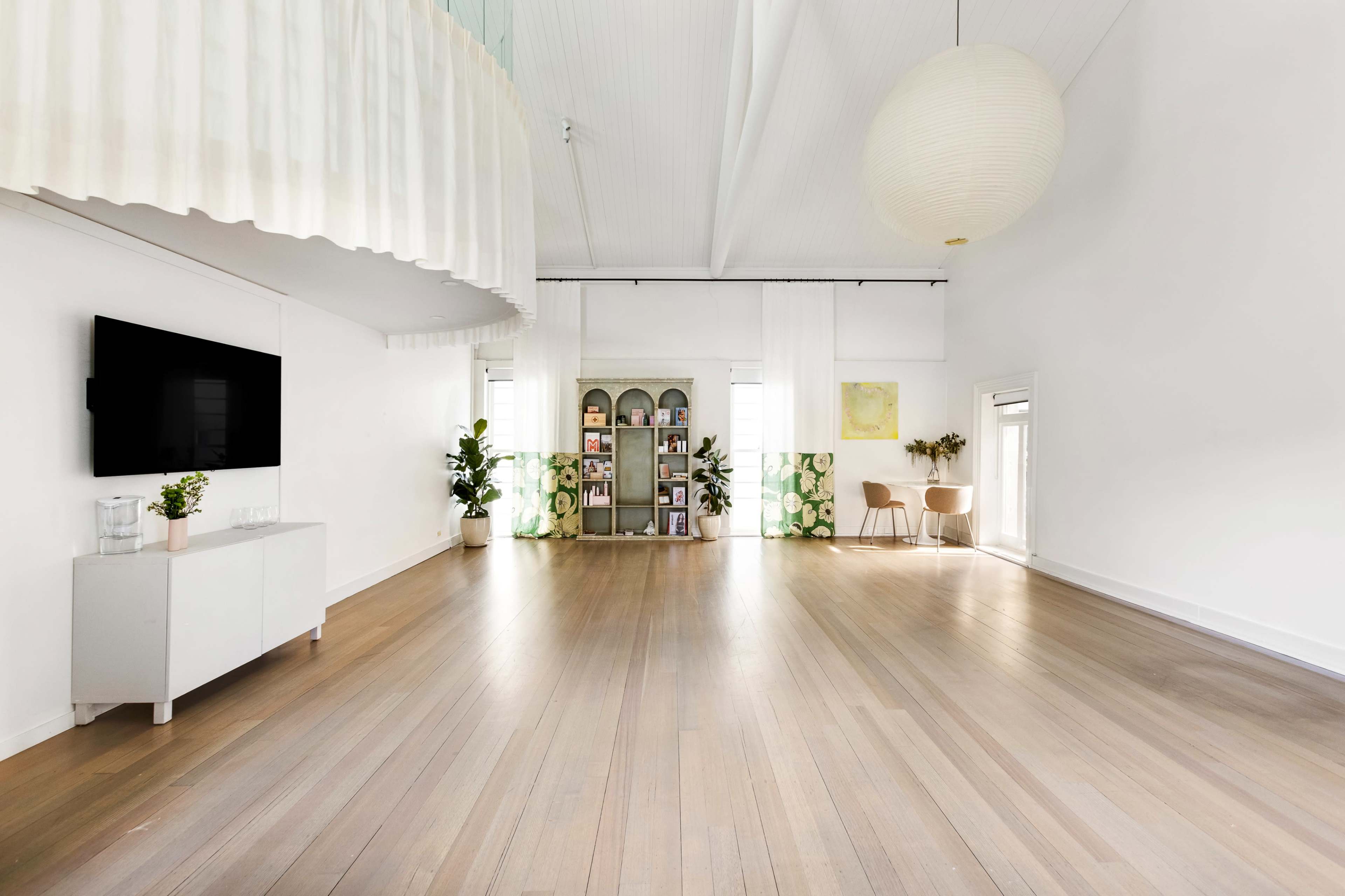 The image shows a bright, open room with wooden floors, a television mounted on the wall, and a shelf filled with books and decorative items in the background.