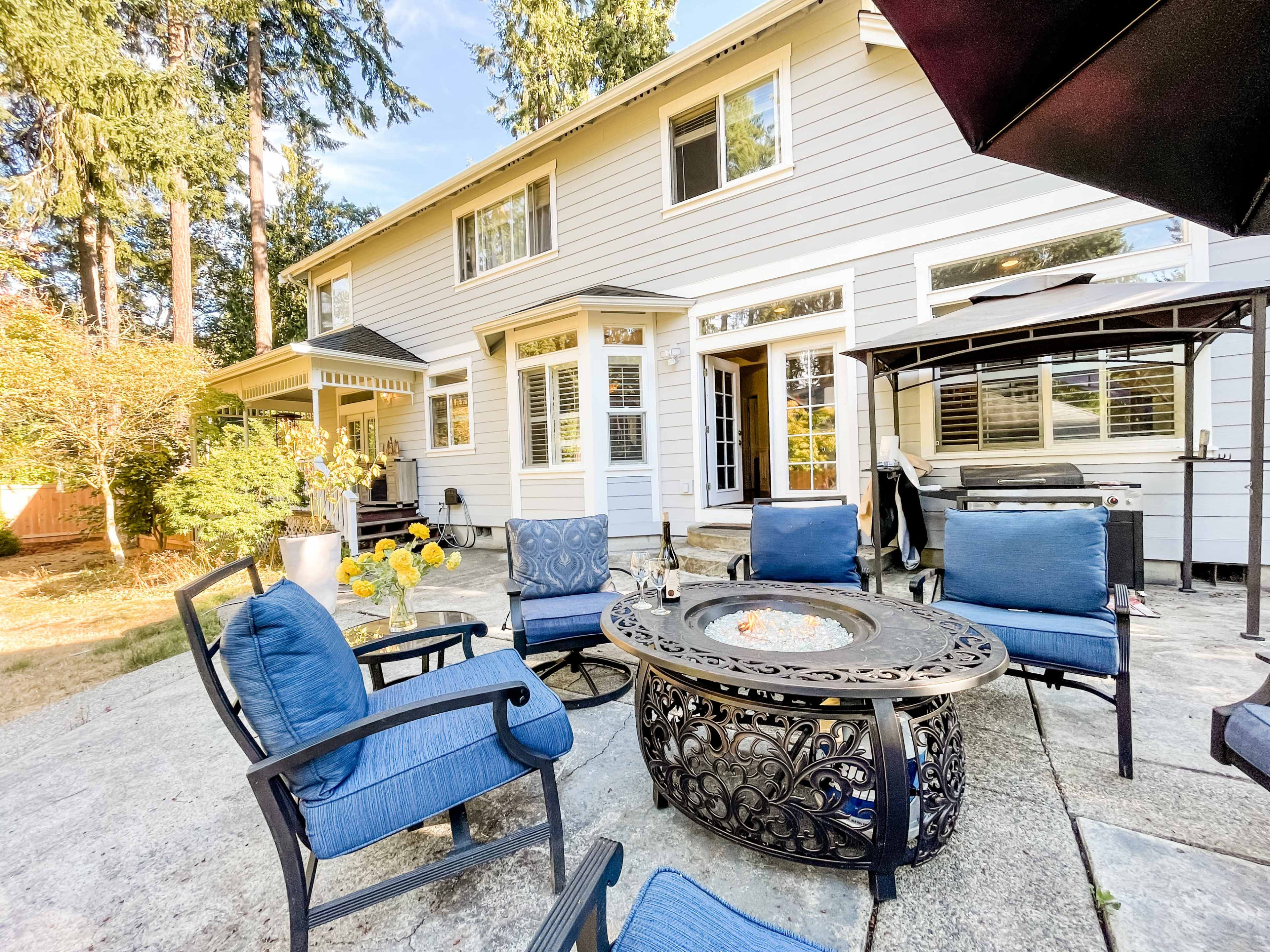 A backyard patio area featuring a fire pit surrounded by blue chairs and a house with large windows in the background.