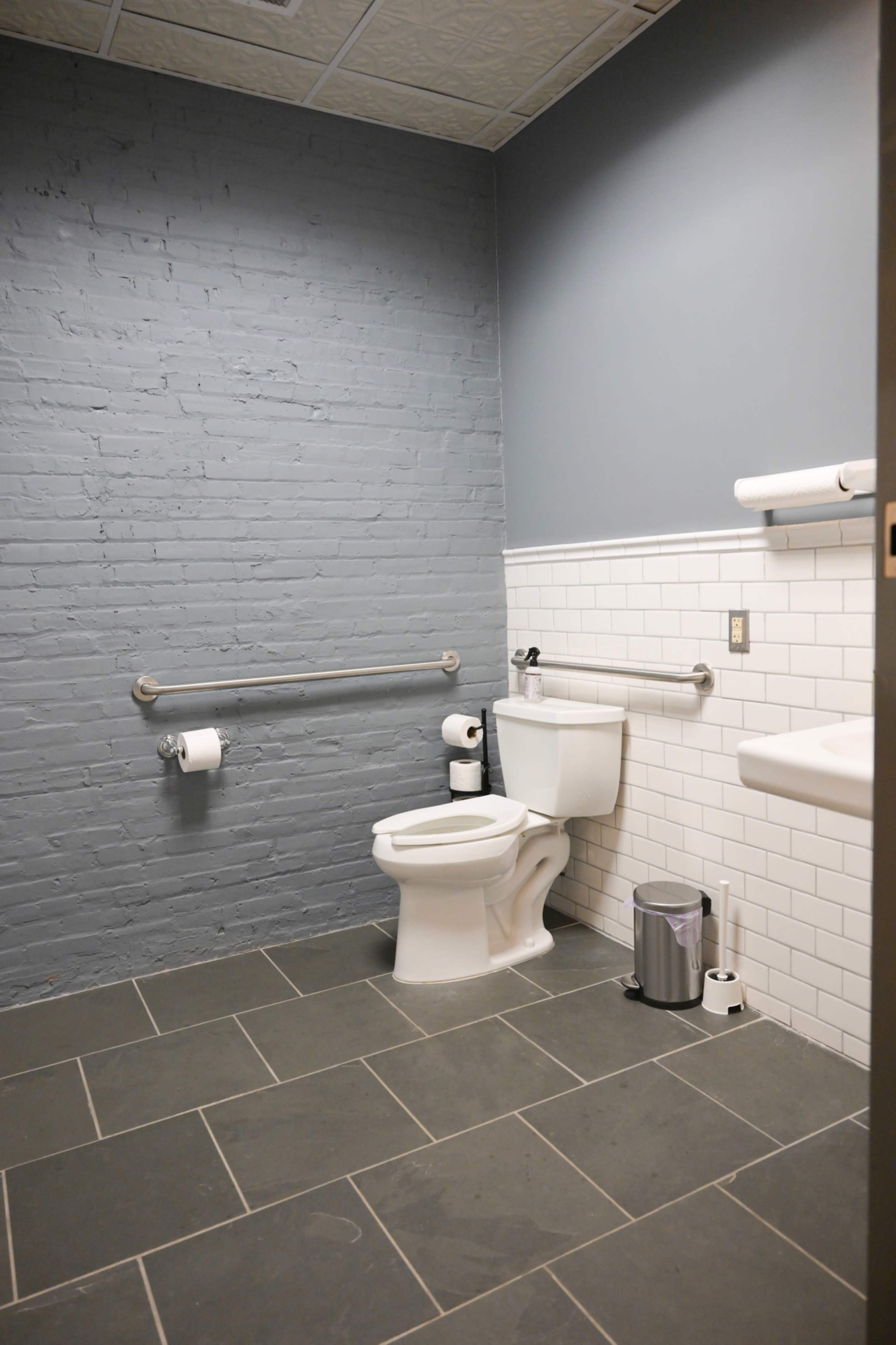 The image shows a simple restroom with a toilet, sink, and gray brick walls, featuring white tiled accents and grab bars.