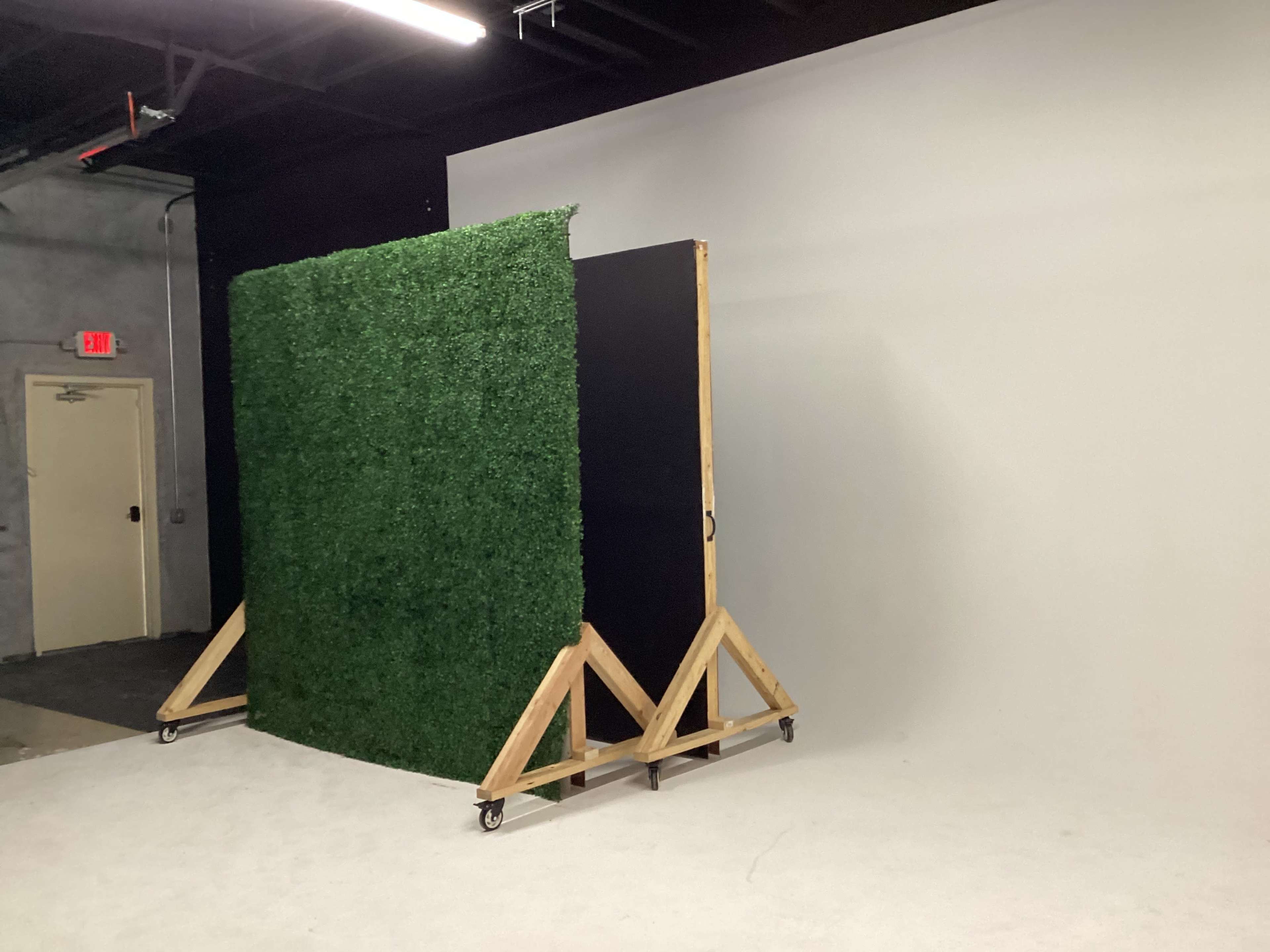 A green artificial grass wall is positioned on a wooden frame with wheels against a plain gray backdrop in a studio space.