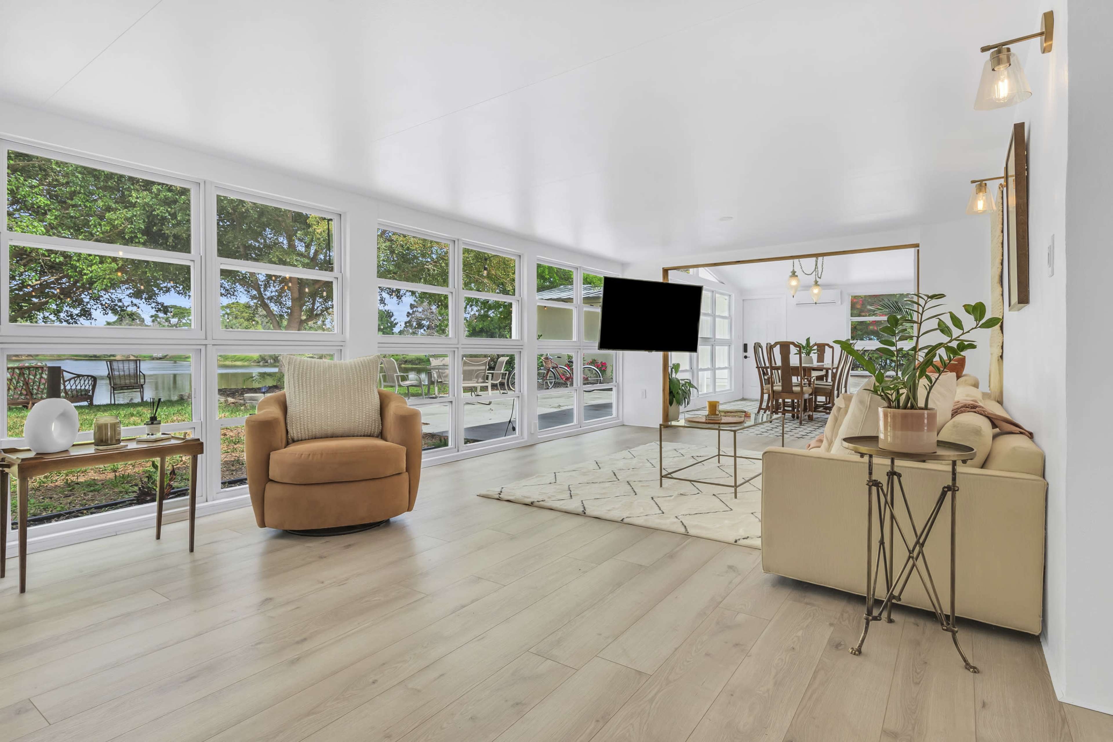 A bright and spacious living area features large windows overlooking a view, with a cozy armchair, a sofa, and a TV mounted on the wall.