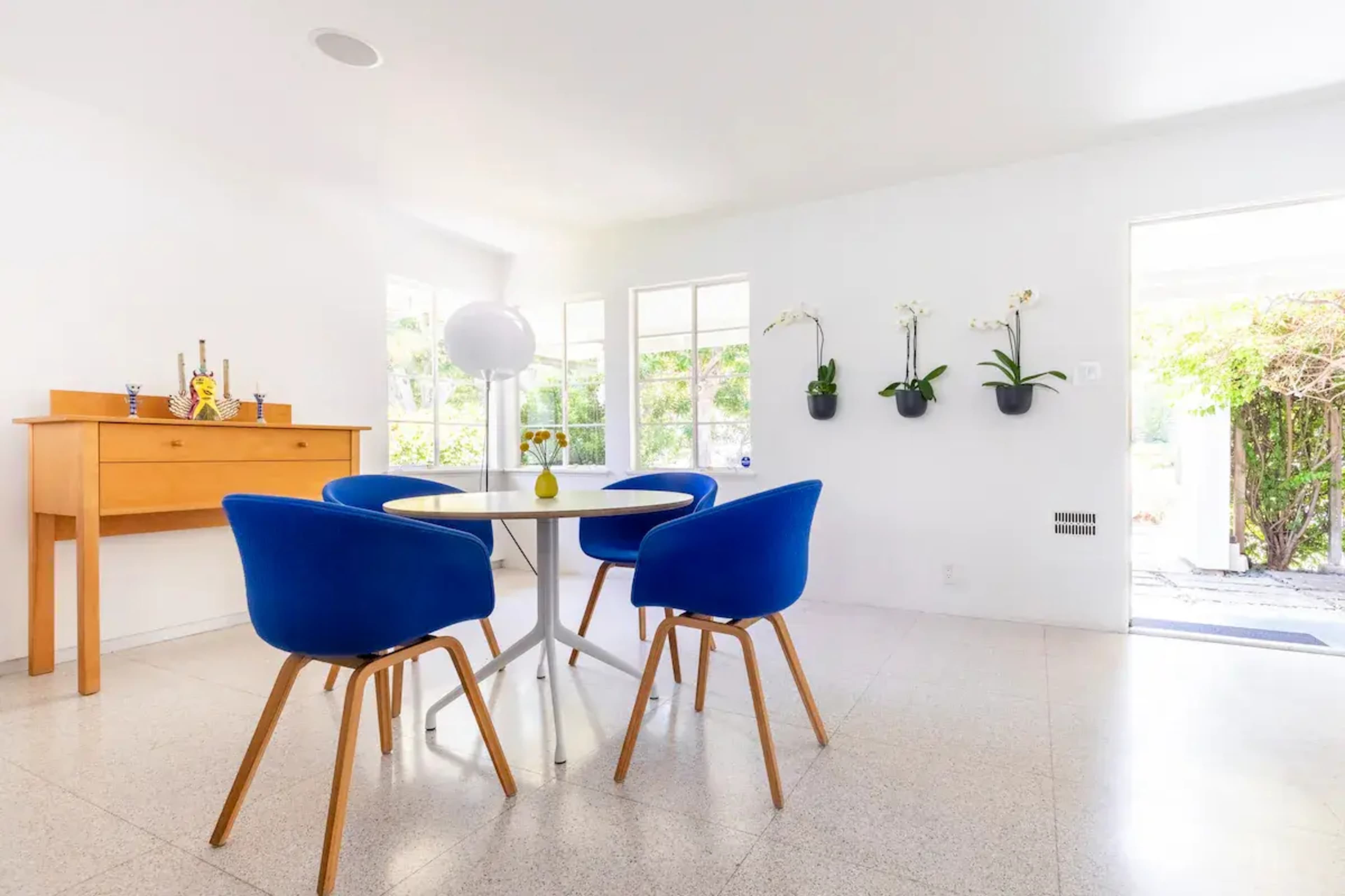 A modern dining area features a round table surrounded by four blue chairs, a wooden sideboard, and light streaming through large windows.