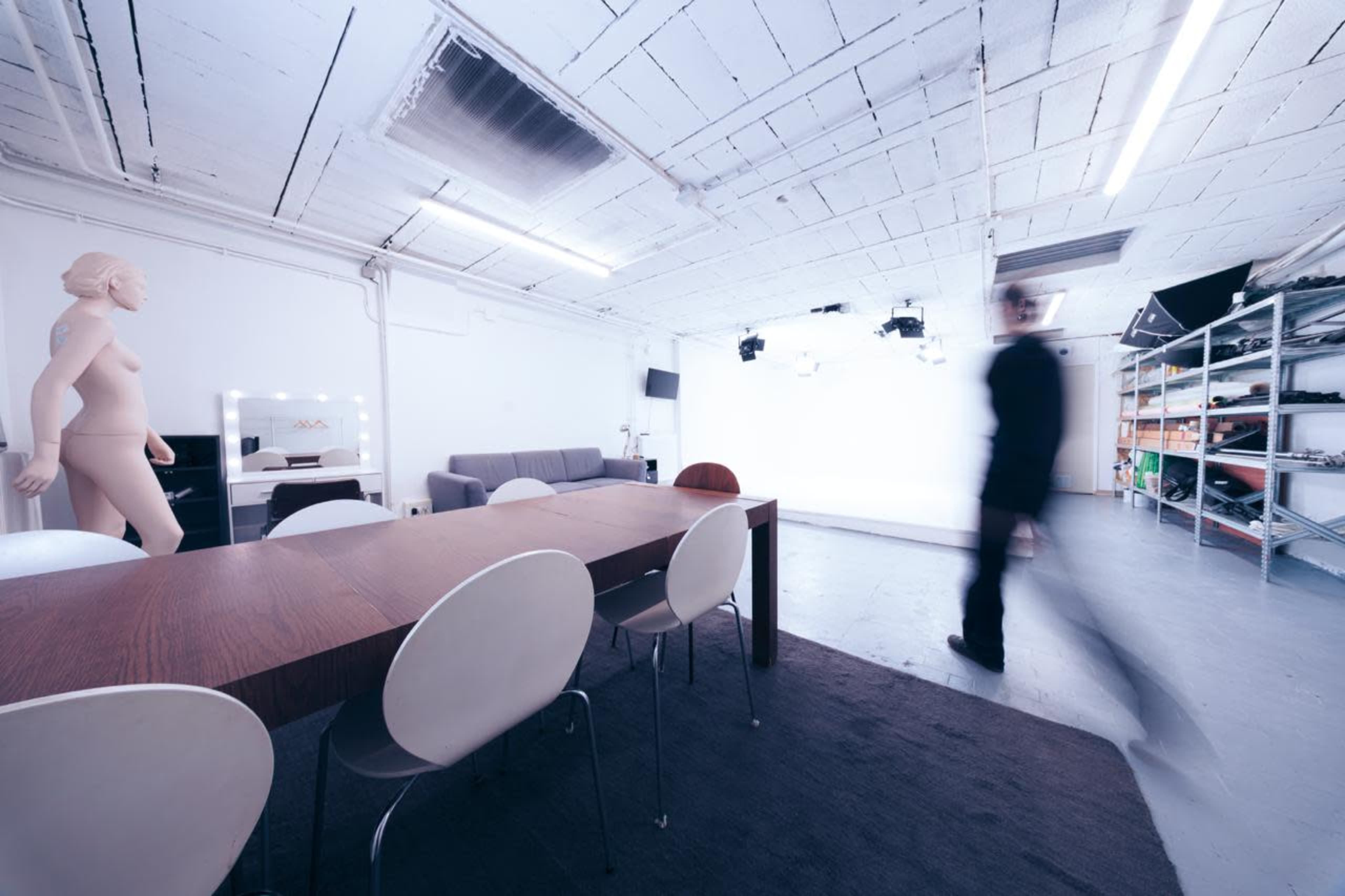 A spacious studio features a wooden table surrounded by white chairs, with a mannequin and a couch in the background.