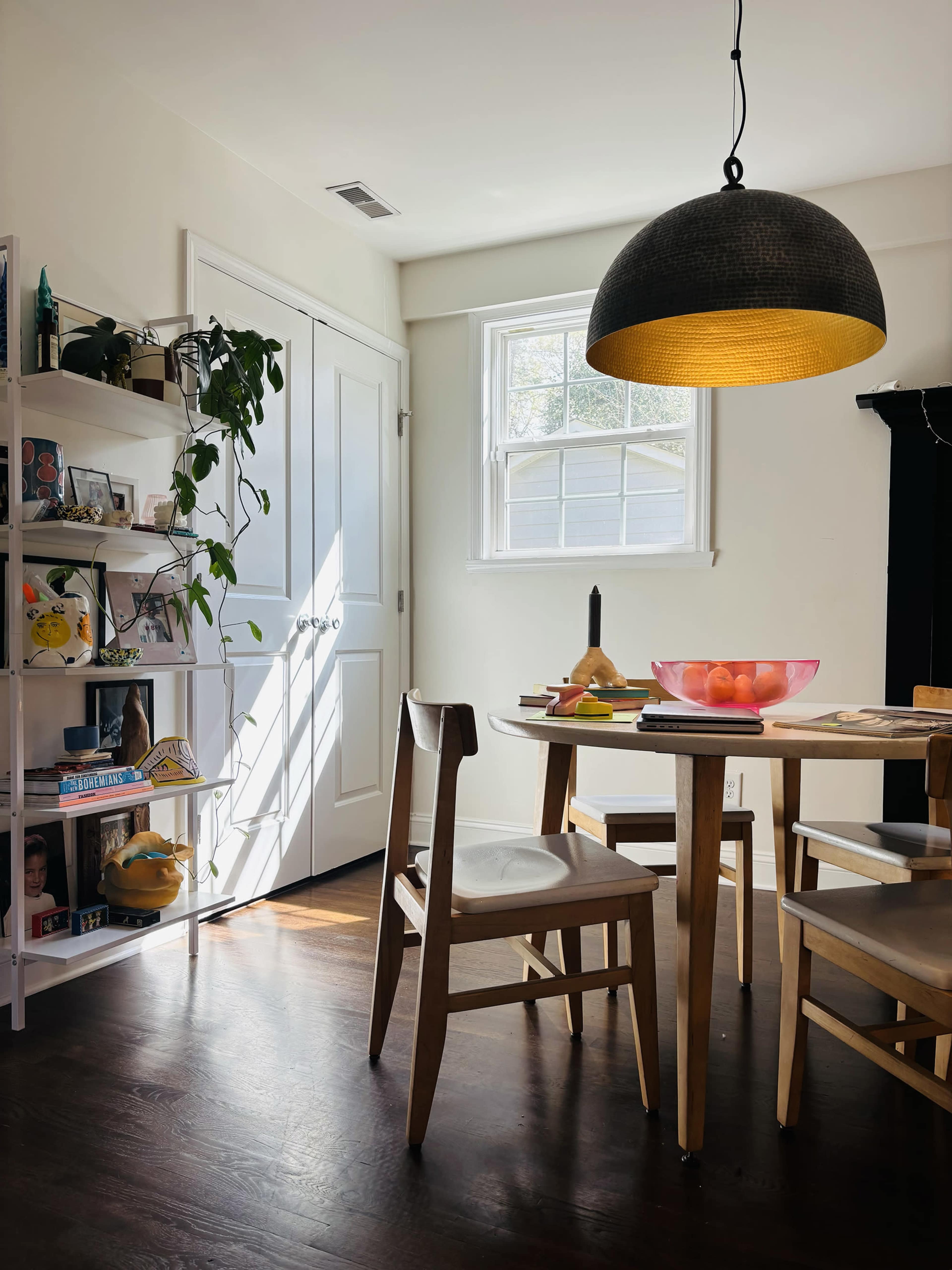 A dining area features a round table with a bowl of fruit, surrounded by wooden chairs, and is illuminated by a pendant light, with a bookshelf and a window in the background.