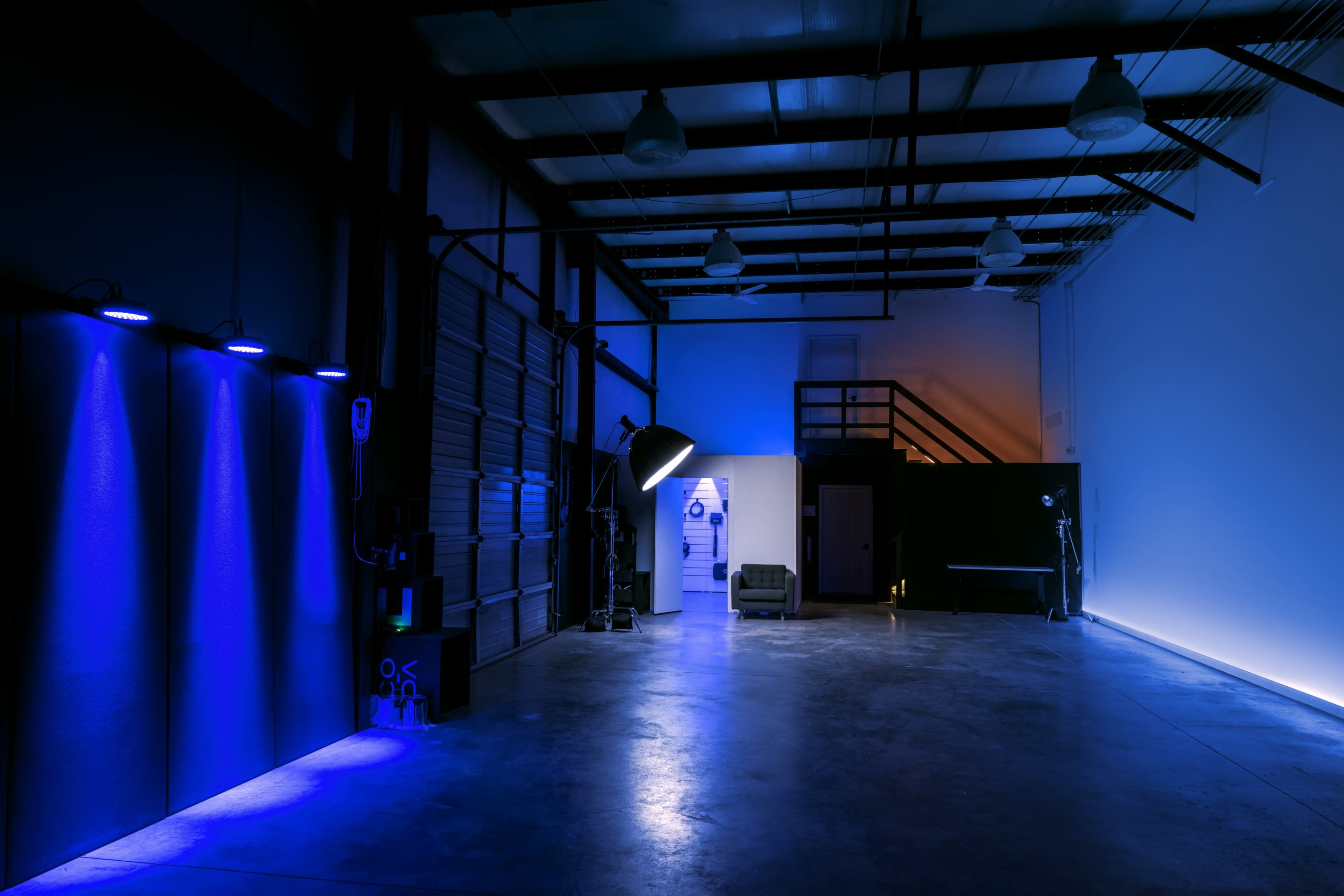 A spacious, dimly lit industrial studio featuring blue lighting, a couch, and various photographic equipment.