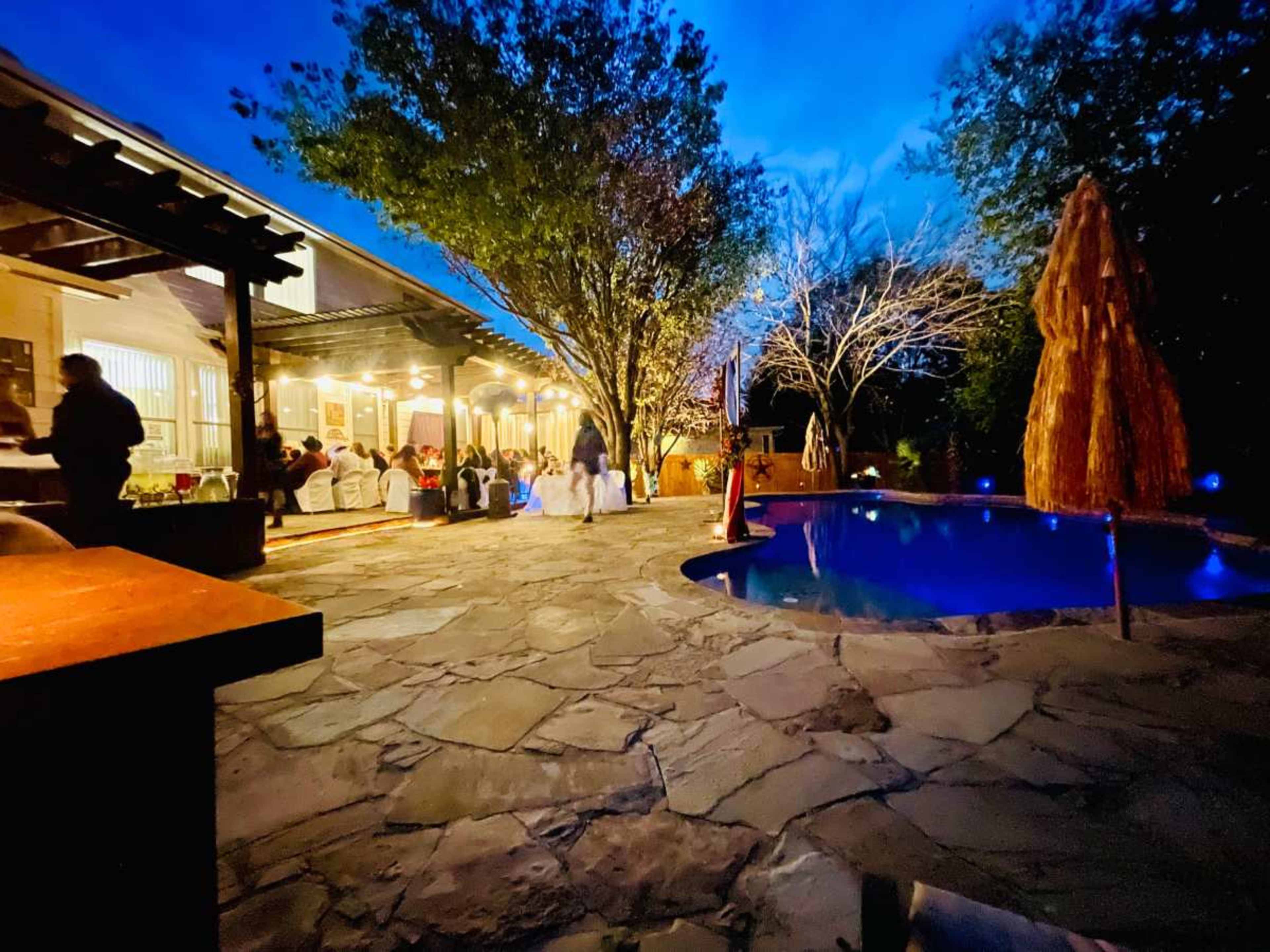 A cozy outdoor gathering is taking place near a pool, with people mingling under trees and ambient lighting at dusk.