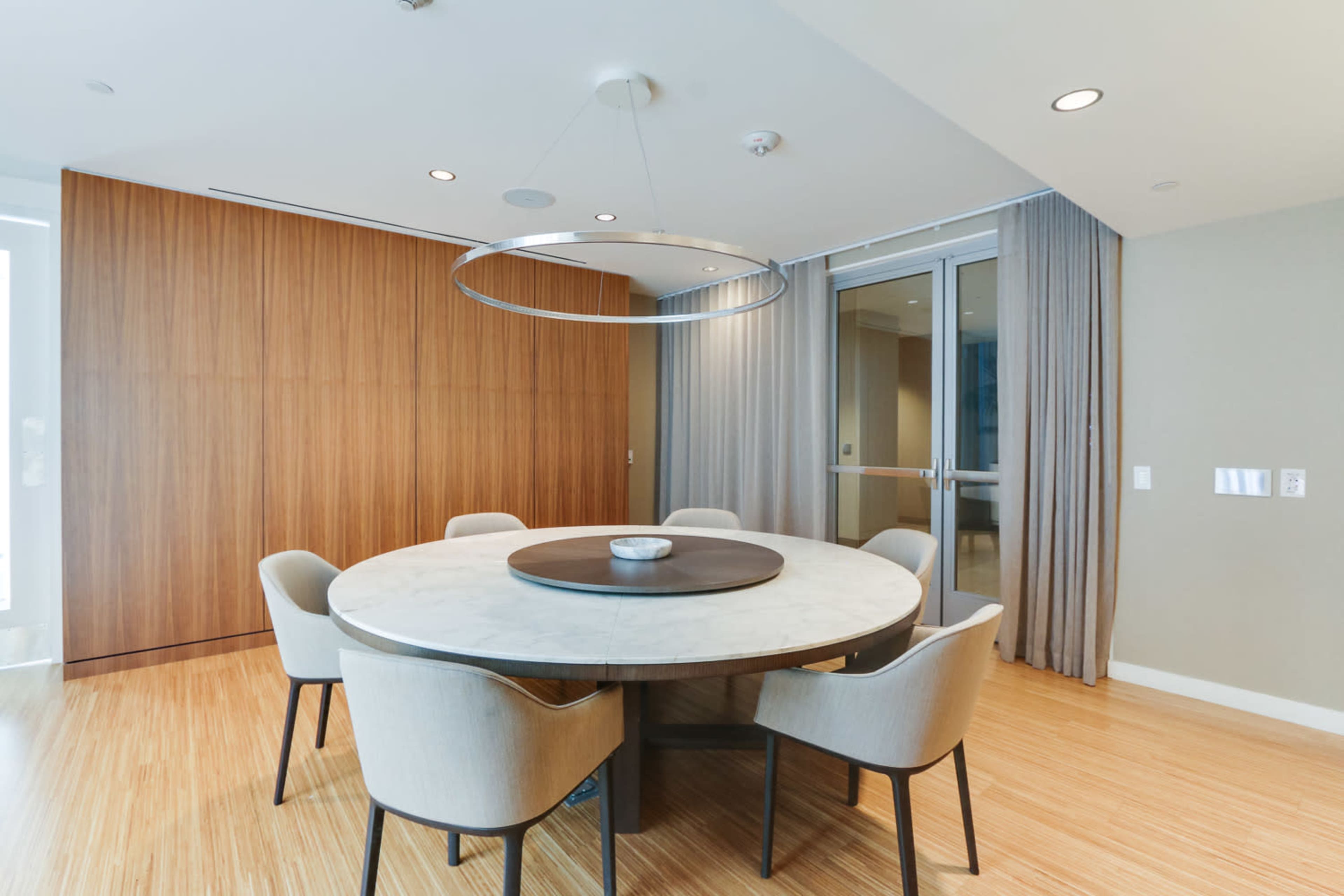 A modern dining area featuring a round table surrounded by six upholstered chairs, illuminated by a circular pendant light.