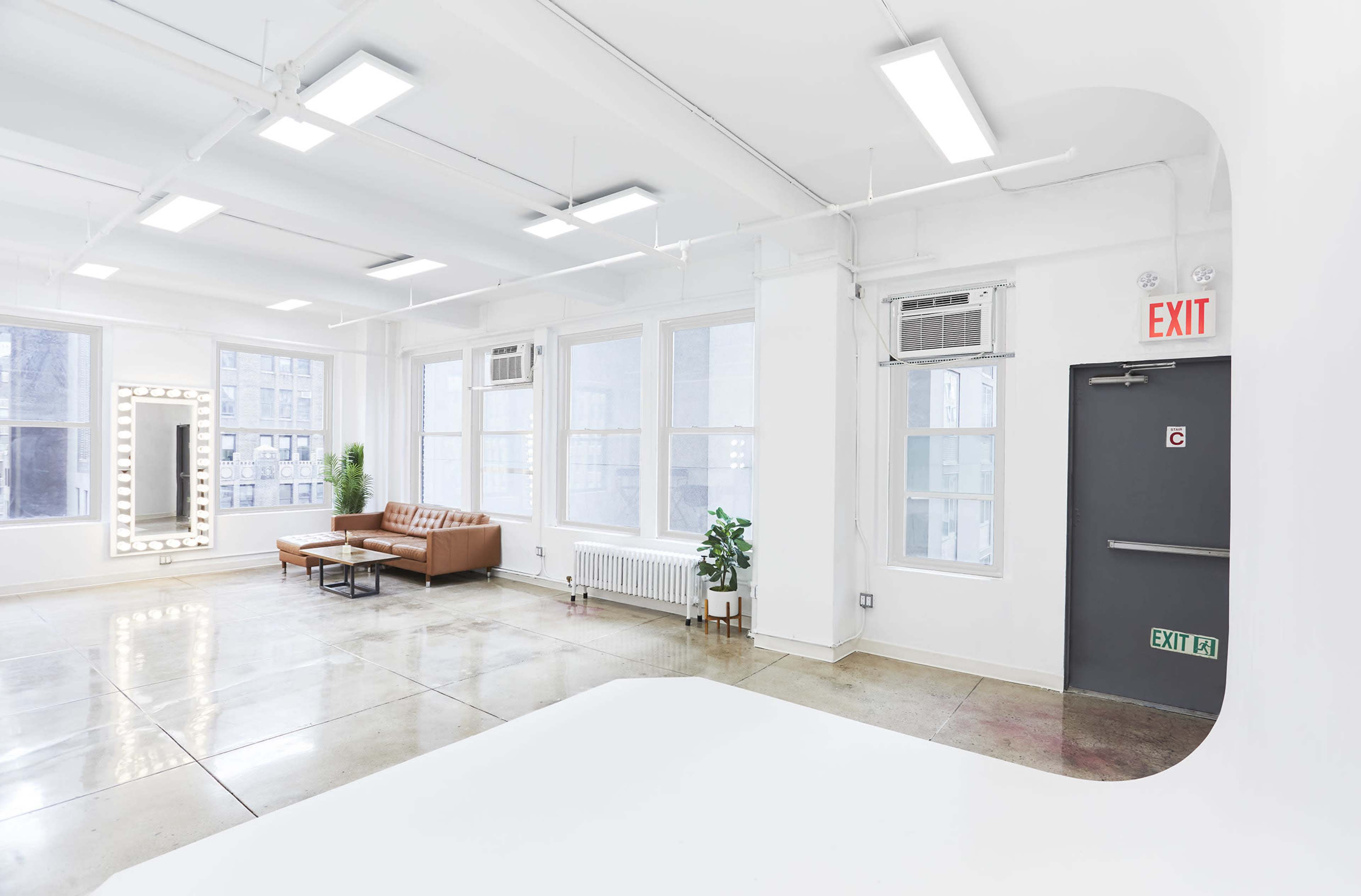 The image shows a bright, spacious room with large windows, a brown couch, a mirrored wall, and an exit door.