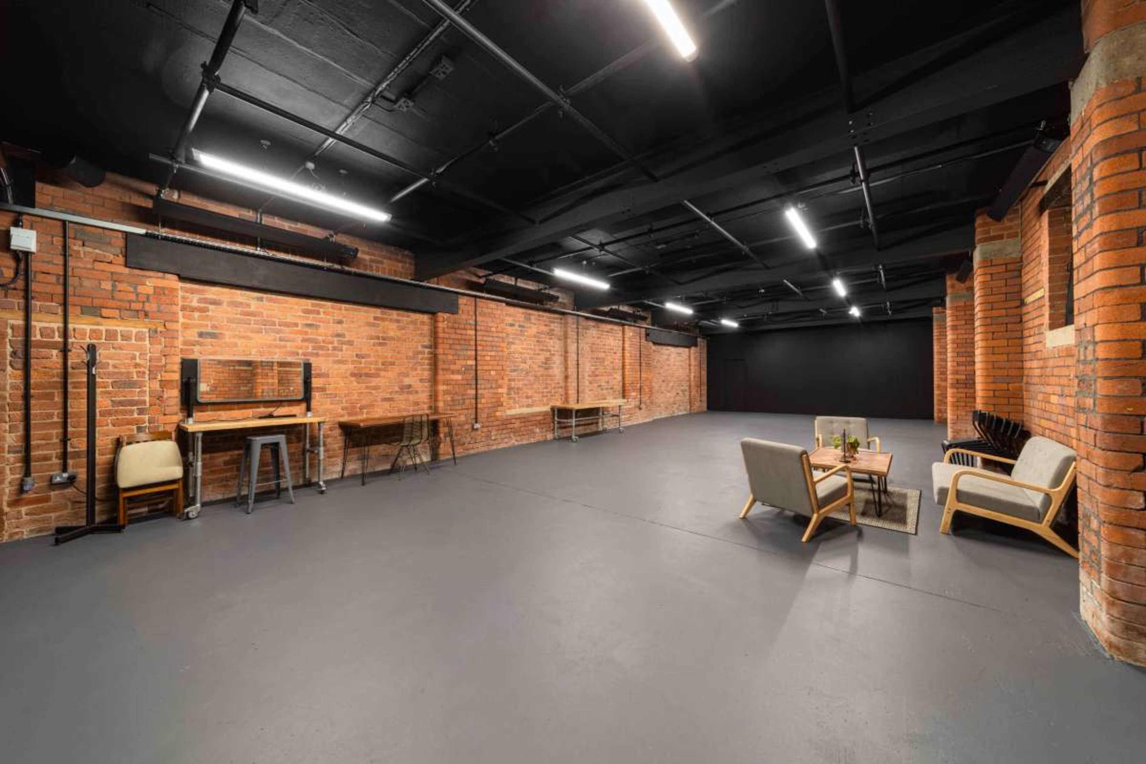 The image shows a spacious industrial-style room with exposed brick walls, concrete flooring, and minimal furniture, including a table and seating area in one corner.