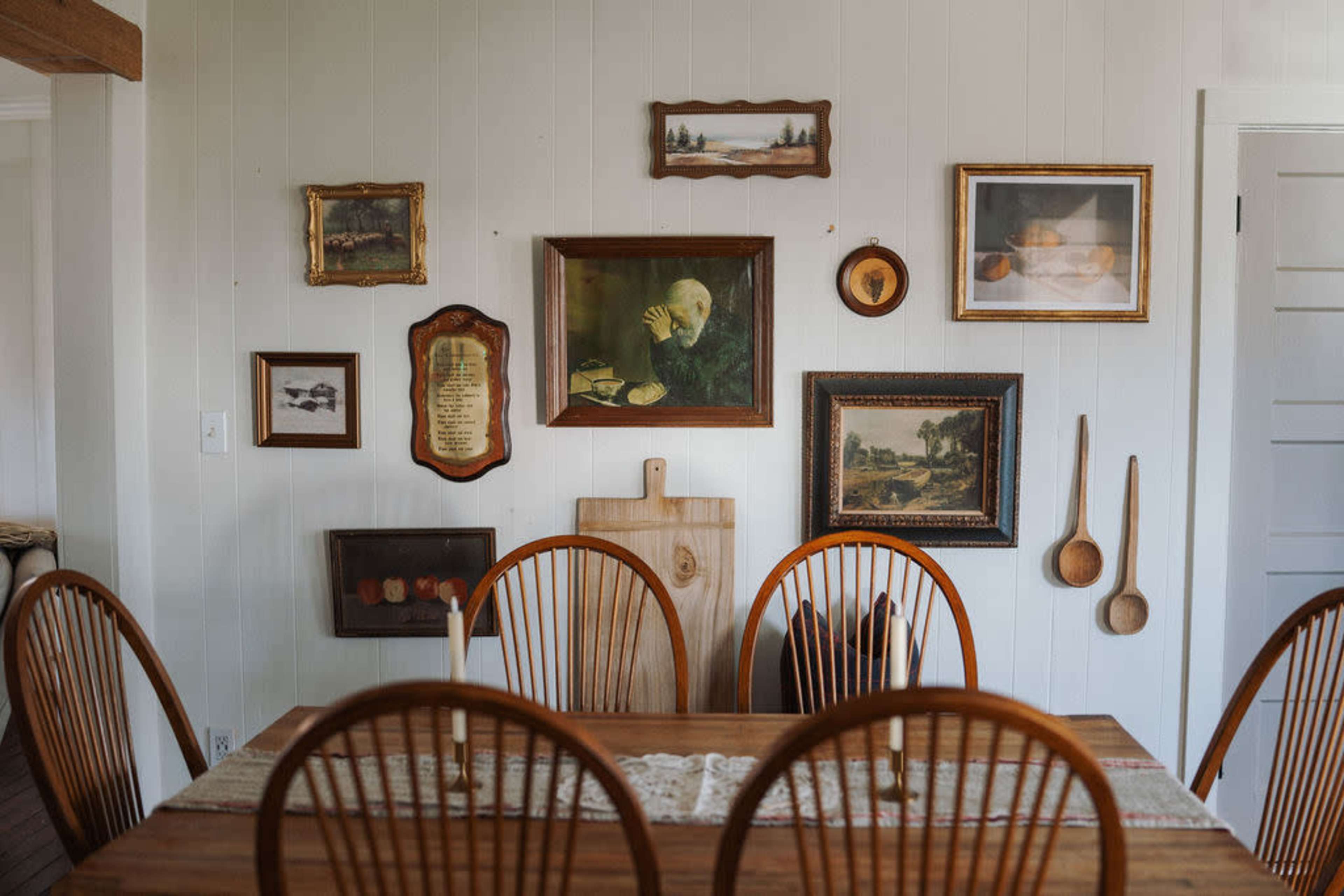 A wooden dining table with several chairs is set against a white wall adorned with various framed paintings and plaques.