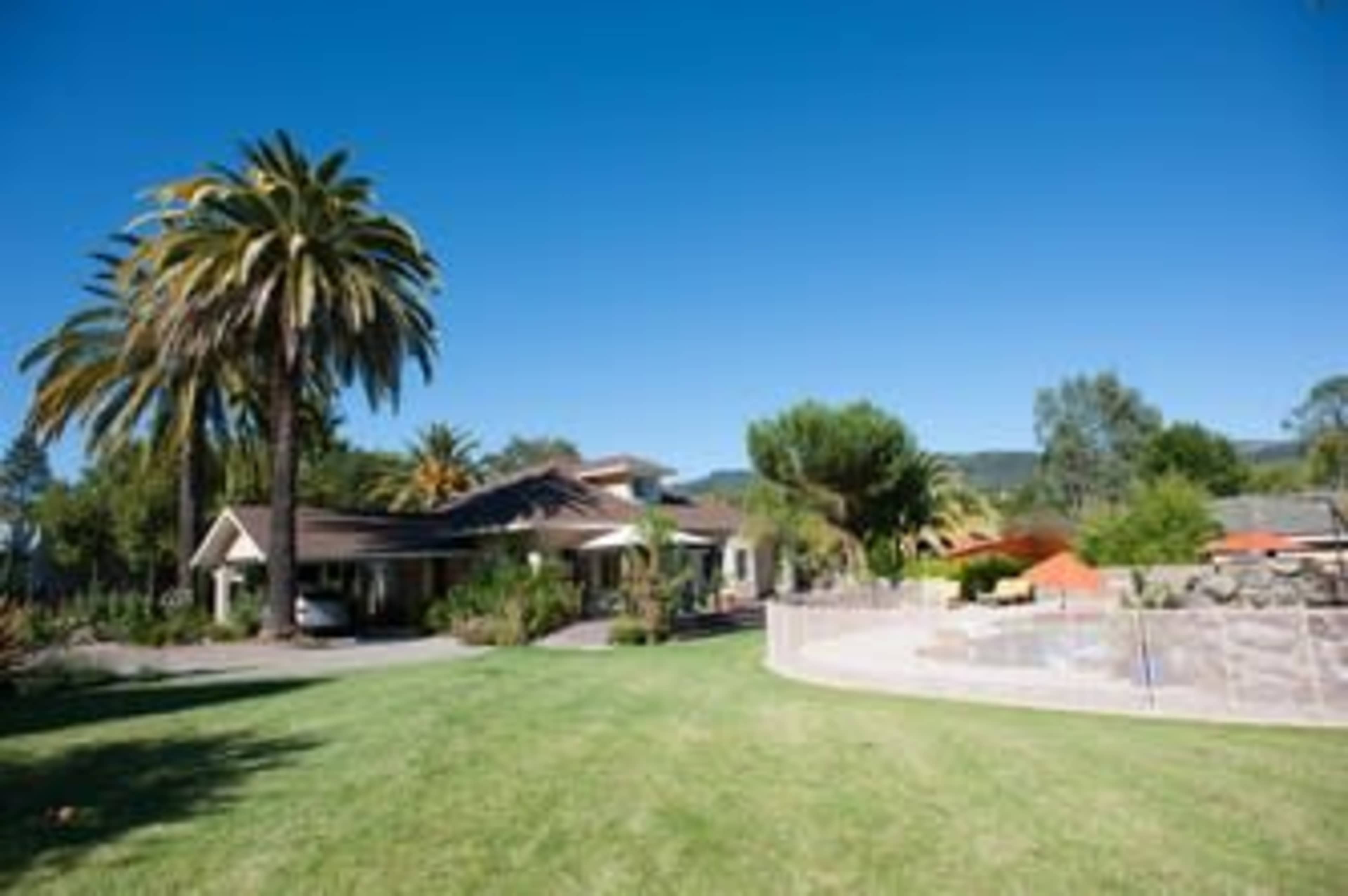 A large house with a landscaped yard and palm trees is situated next to a pool area in a sunny setting.