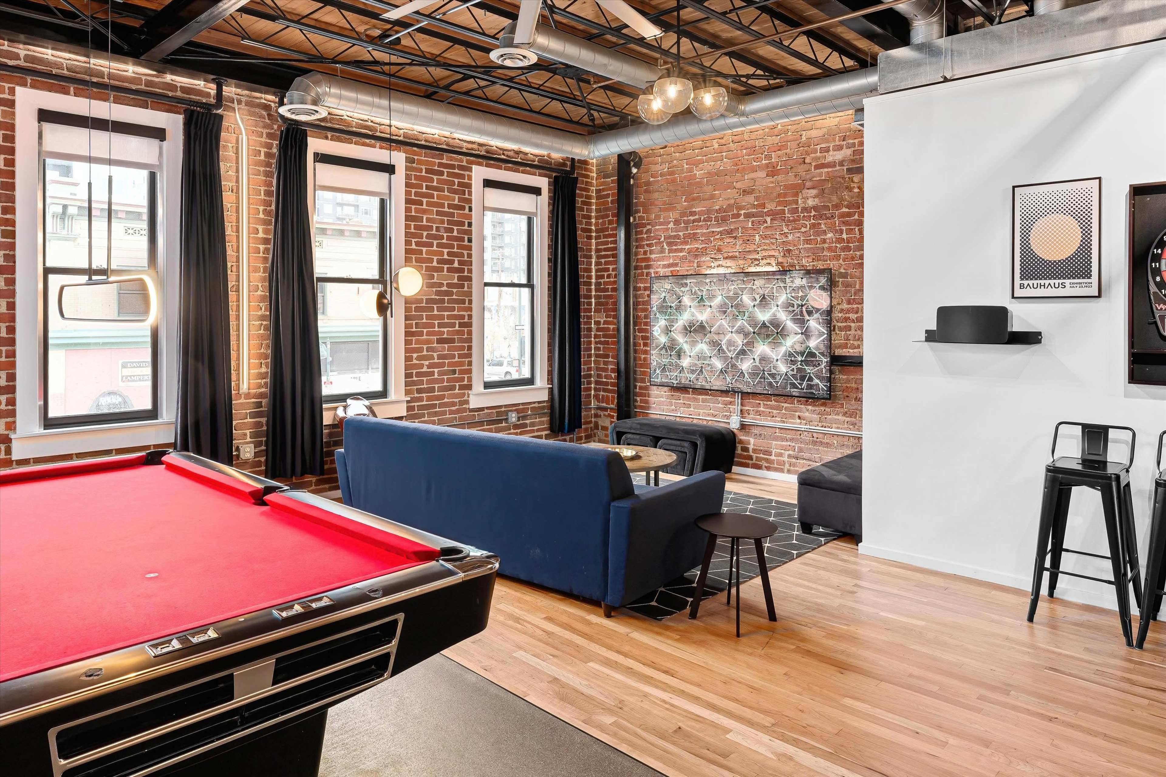 A living space with a red pool table in the foreground, a blue sofa and a coffee table in the center, and large windows with black curtains along the brick wall.