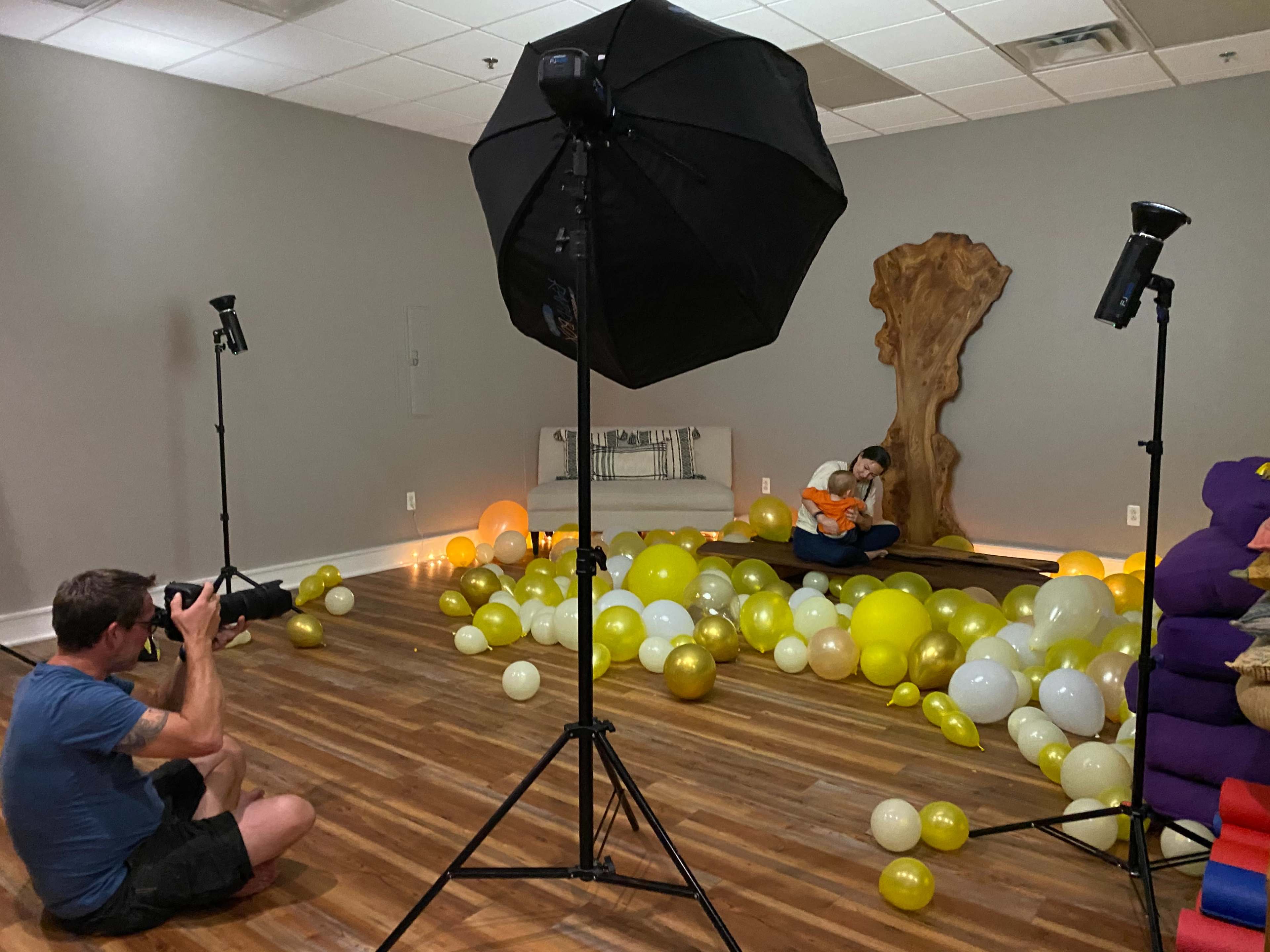 A photographer captures a child sitting amidst yellow and transparent balloons in a studio setting with softbox lighting.