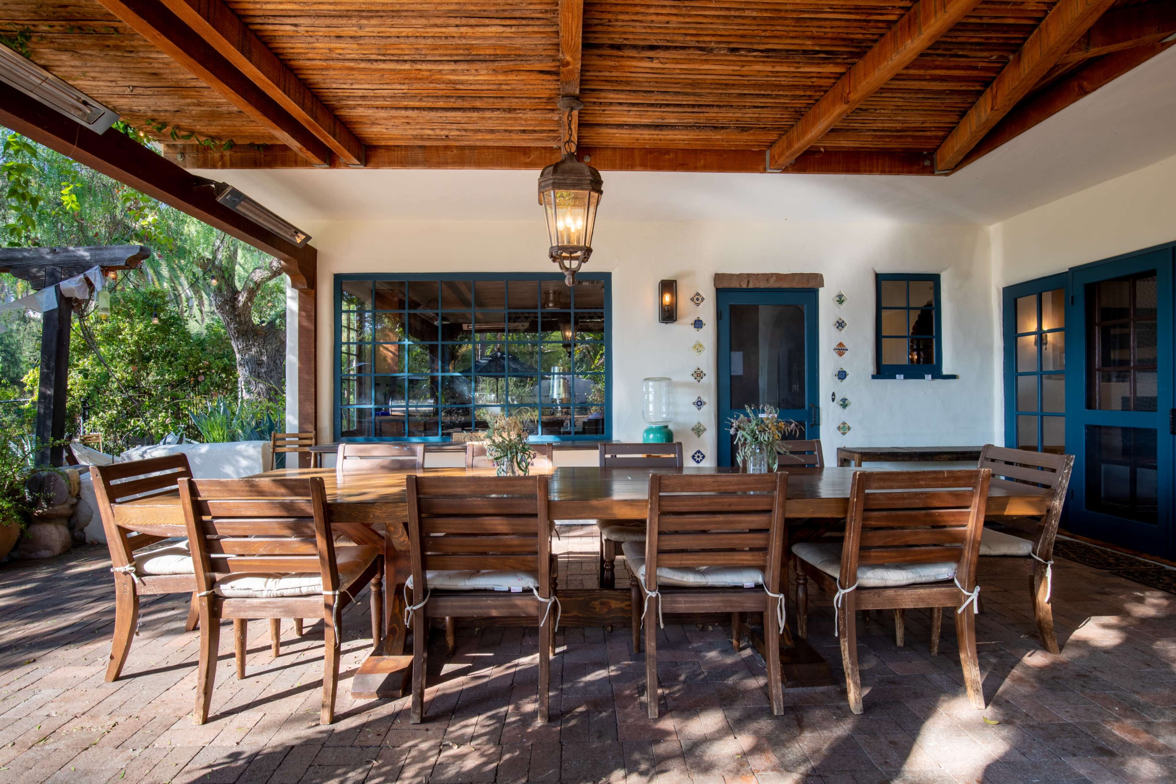 A wooden dining table with matching chairs is set under a covered patio, featuring exposed beams and a lantern hanging overhead.