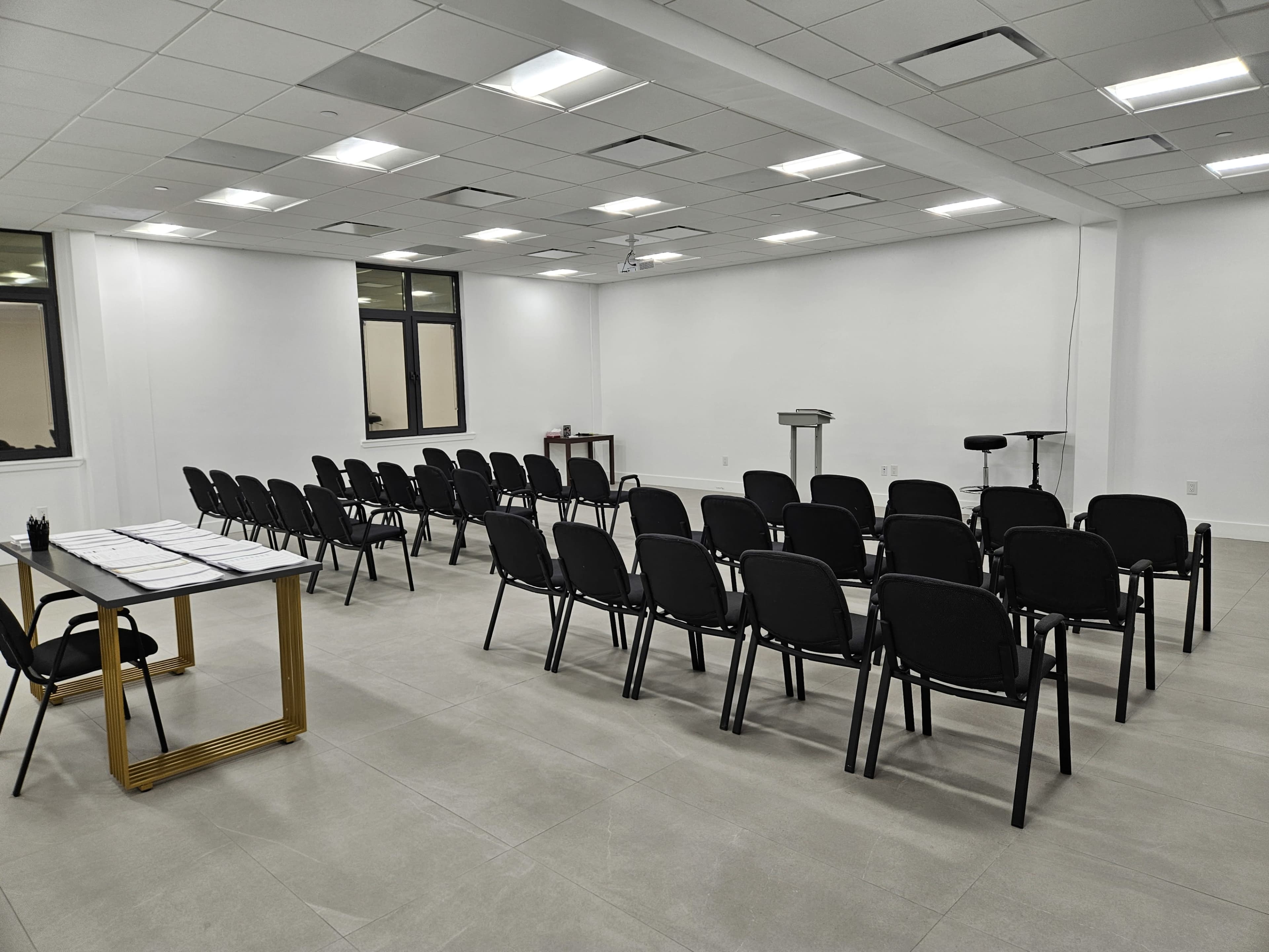 The image shows a sparsely furnished meeting room with rows of black chairs facing a podium and a projector screen on a white wall.