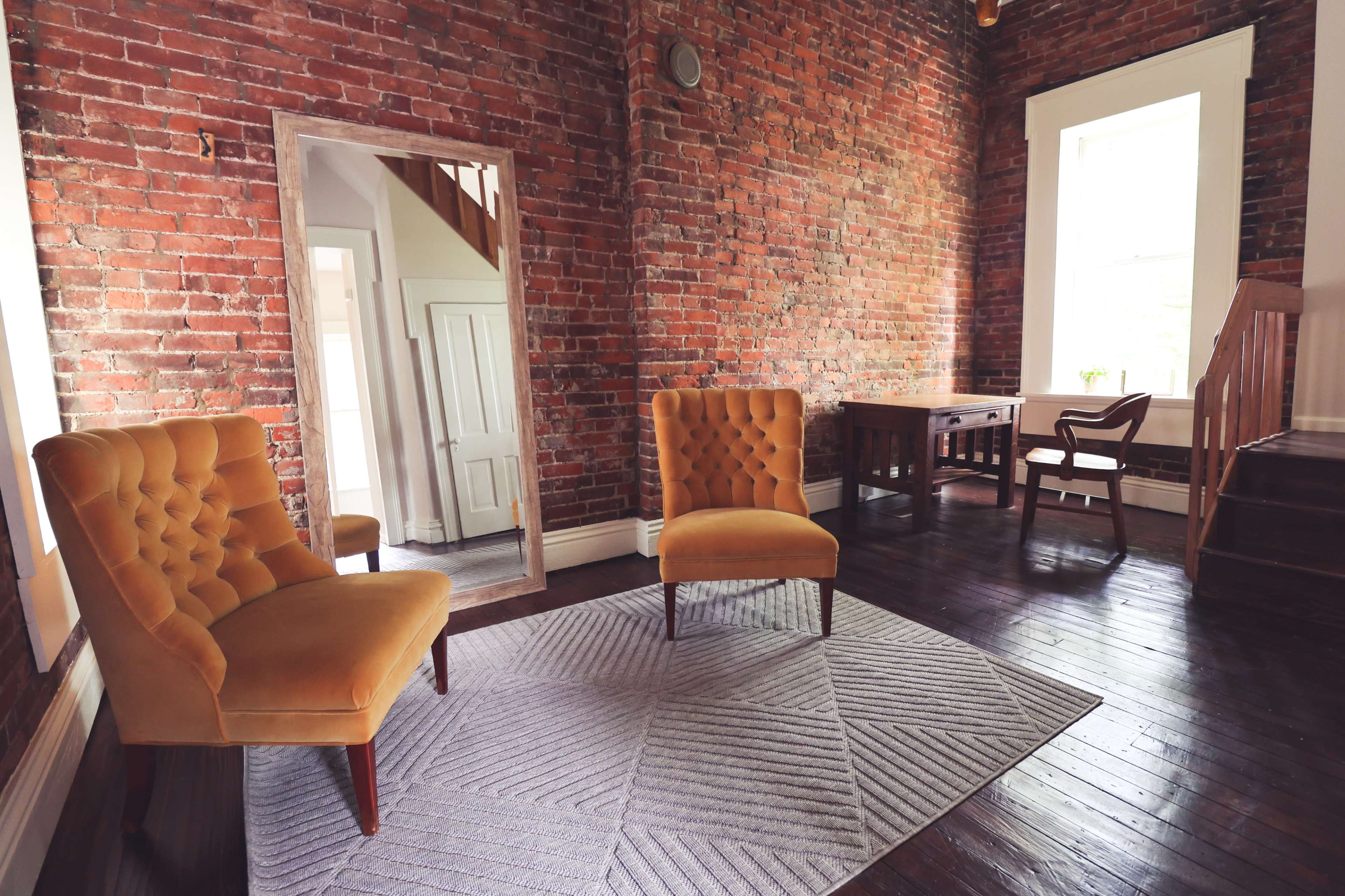 A room with exposed brick walls, two orange armchairs, a patterned rug, and a wooden desk next to a staircase.