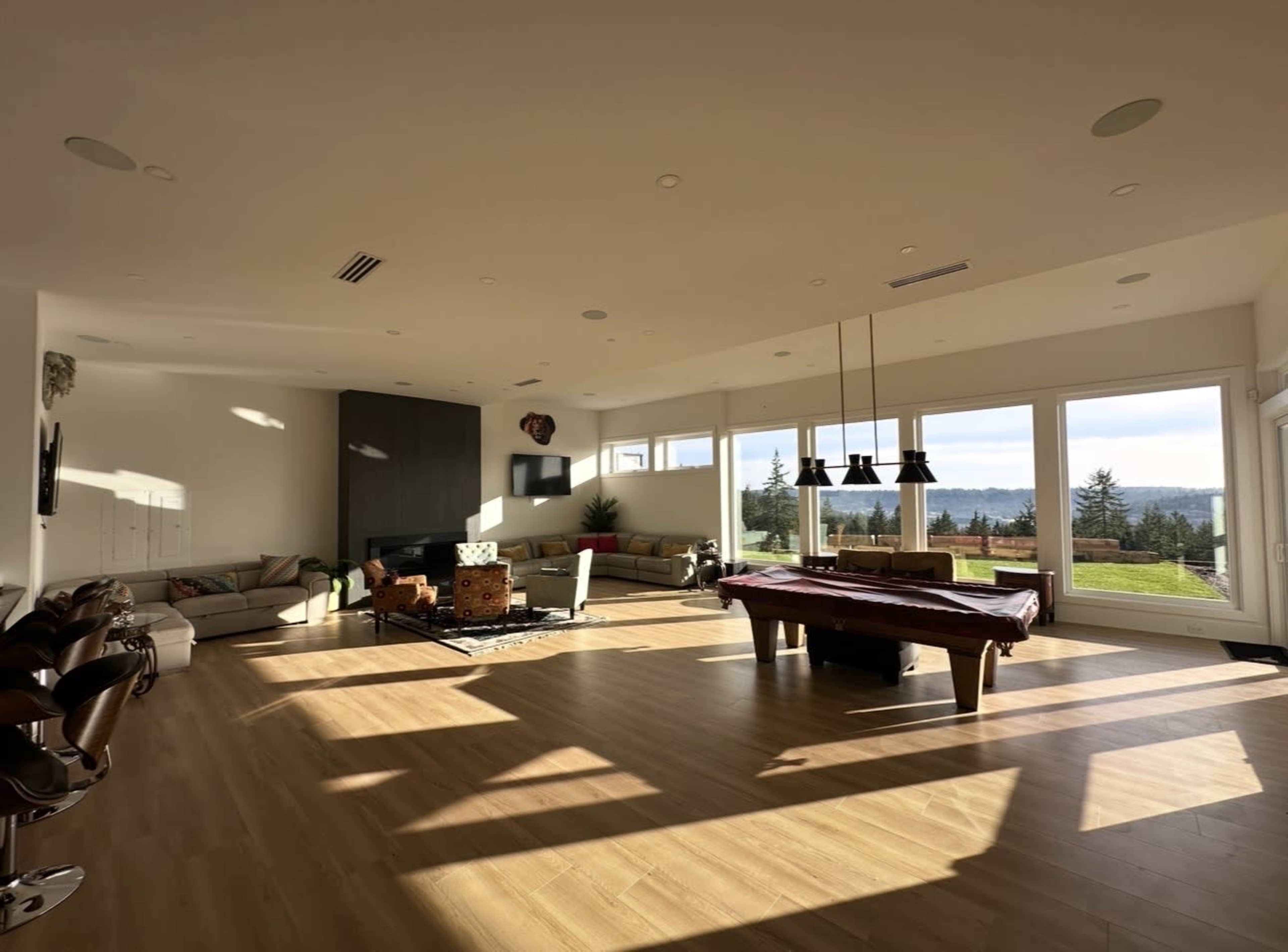 The image depicts a spacious, well-lit living room featuring a pool table, large windows with a view of trees and hills, and modern furnishings.