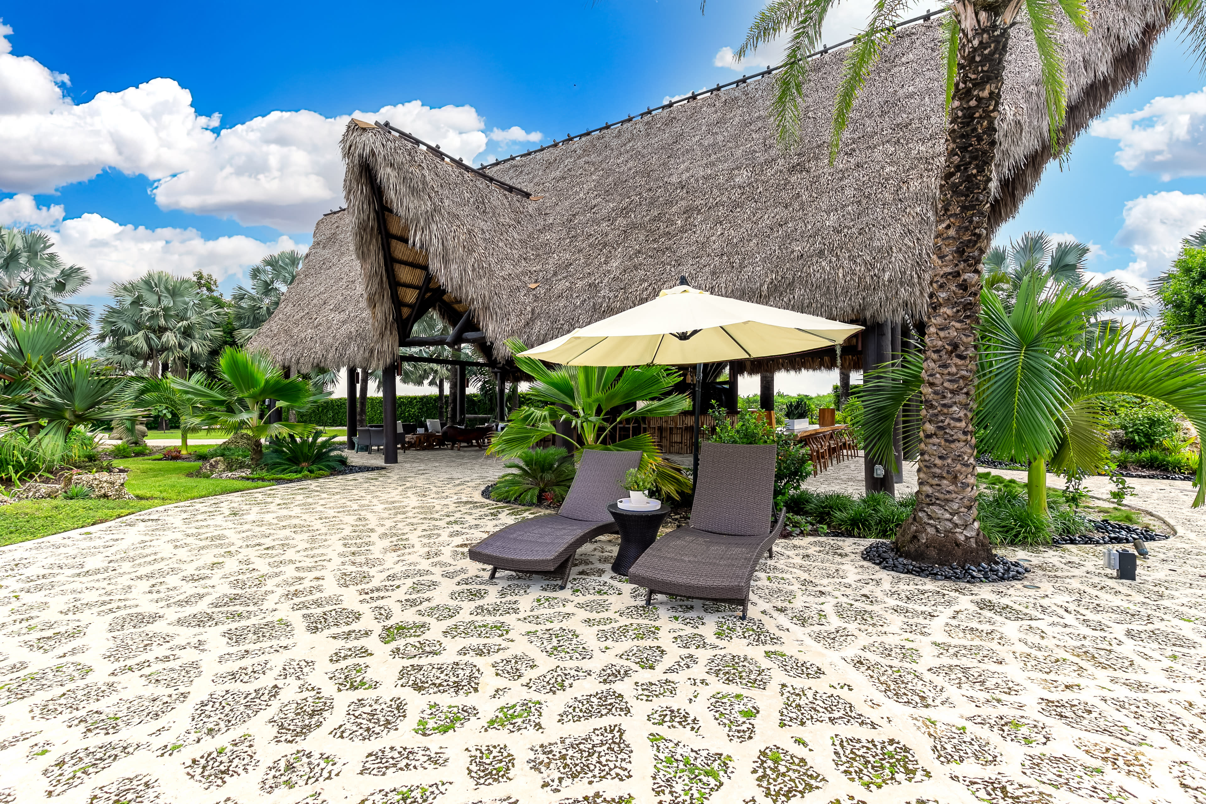 A thatched-roof structure is surrounded by tropical landscaping and lounge chairs beside a large umbrella on a patterned stone patio.