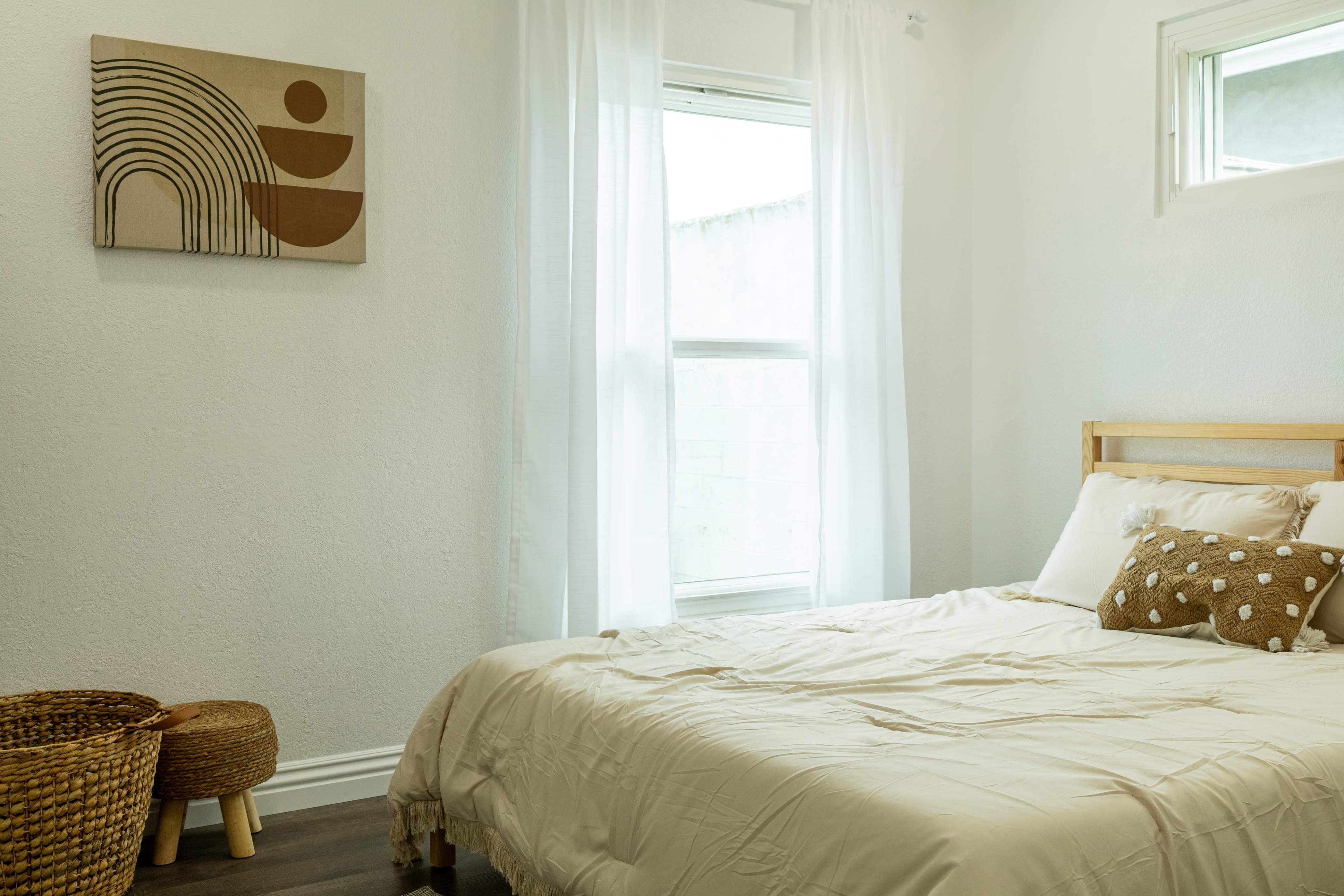A minimalistic bedroom features a bed with light bedding, a decorative pillow, a window dressed with sheer curtains, and a small woven basket beside the bed.
