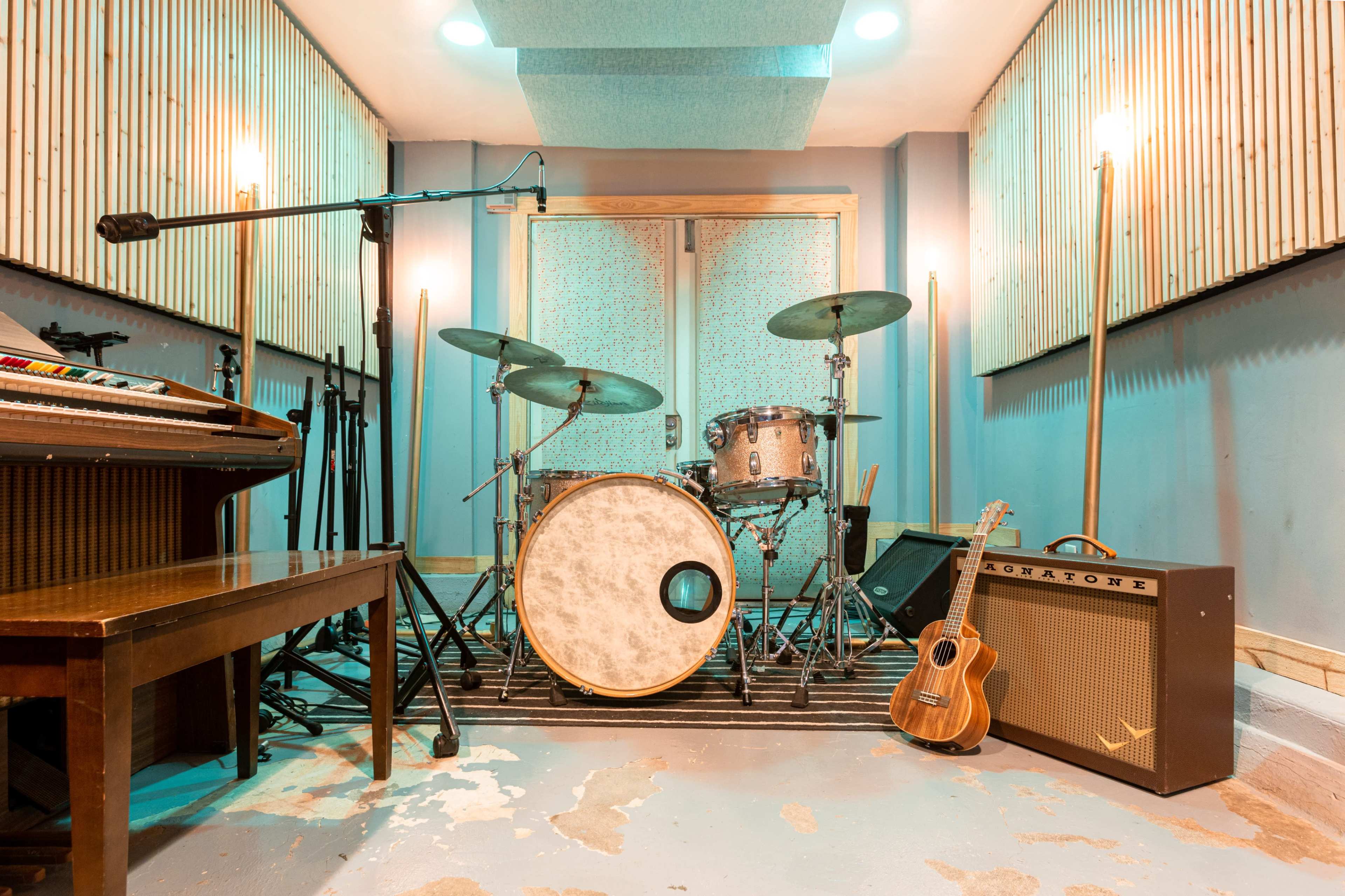 A music studio with a drum set, keyboard, ukulele, and amplifiers arranged in a brightly lit space with wooden wall panels.