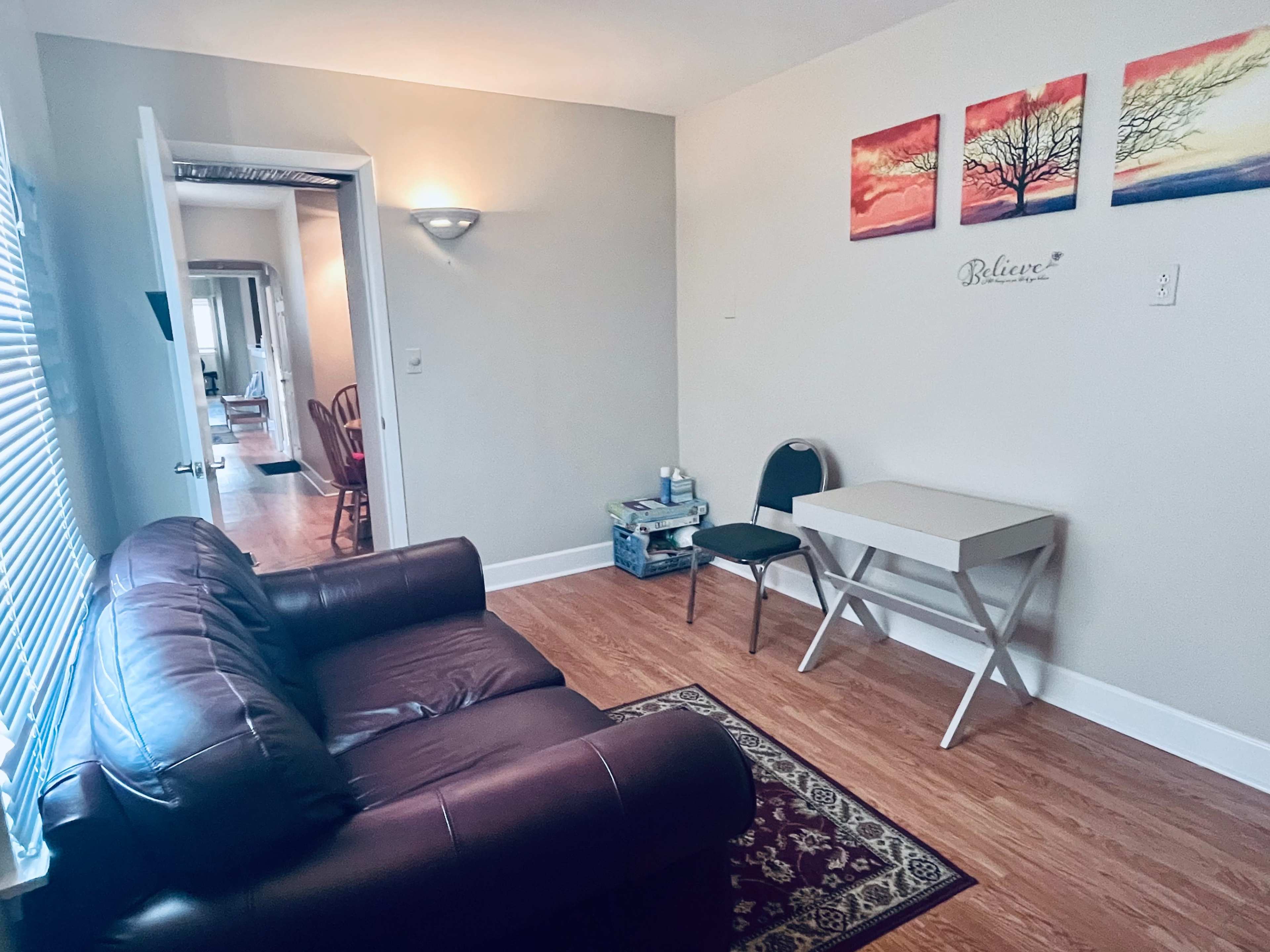 A small room features a brown leather sofa, a table with a chair, and wall decorations depicting trees and the word "Believe."