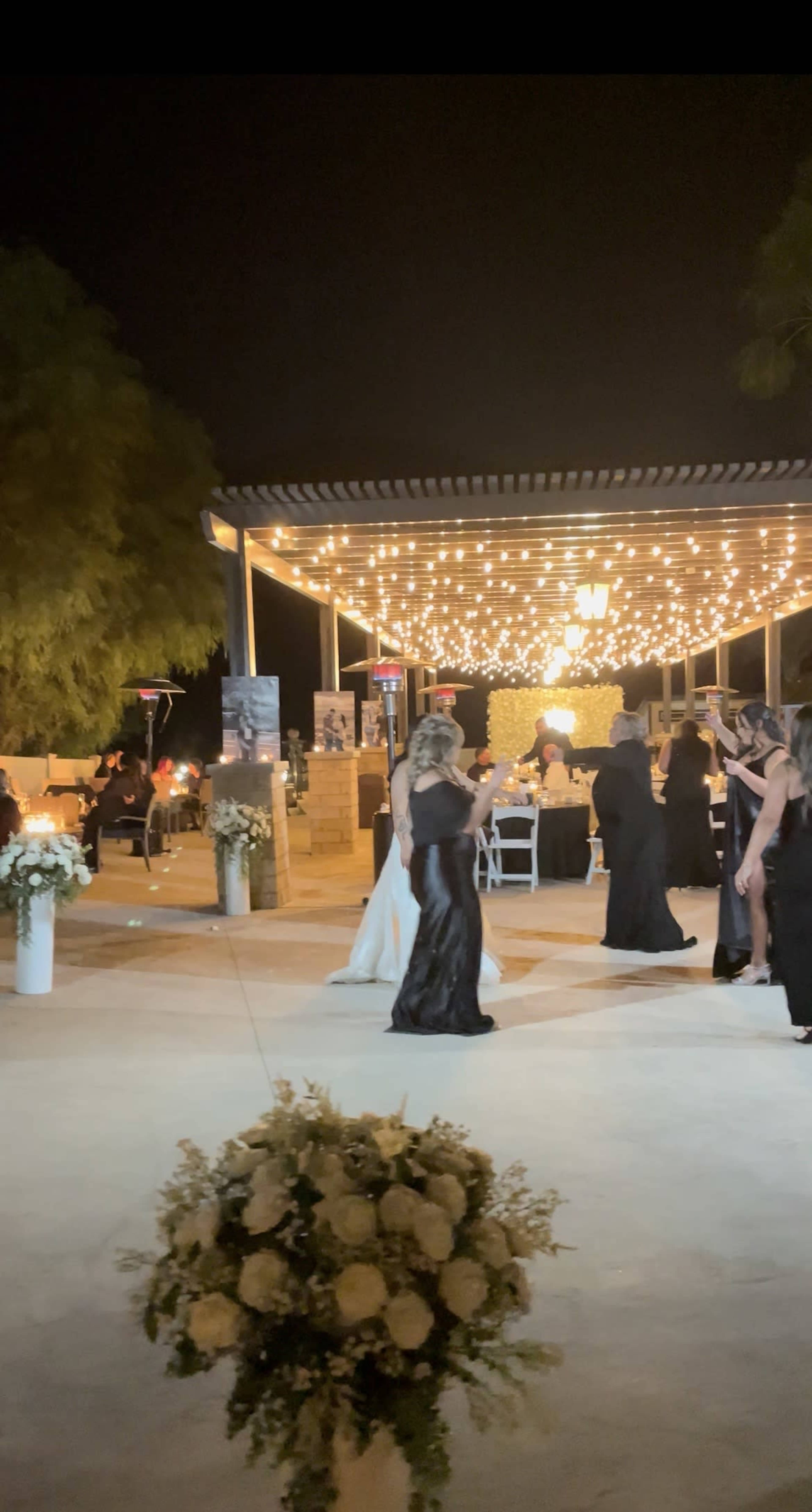 A group of people in formal attire are dancing under string lights at an outdoor event venue.