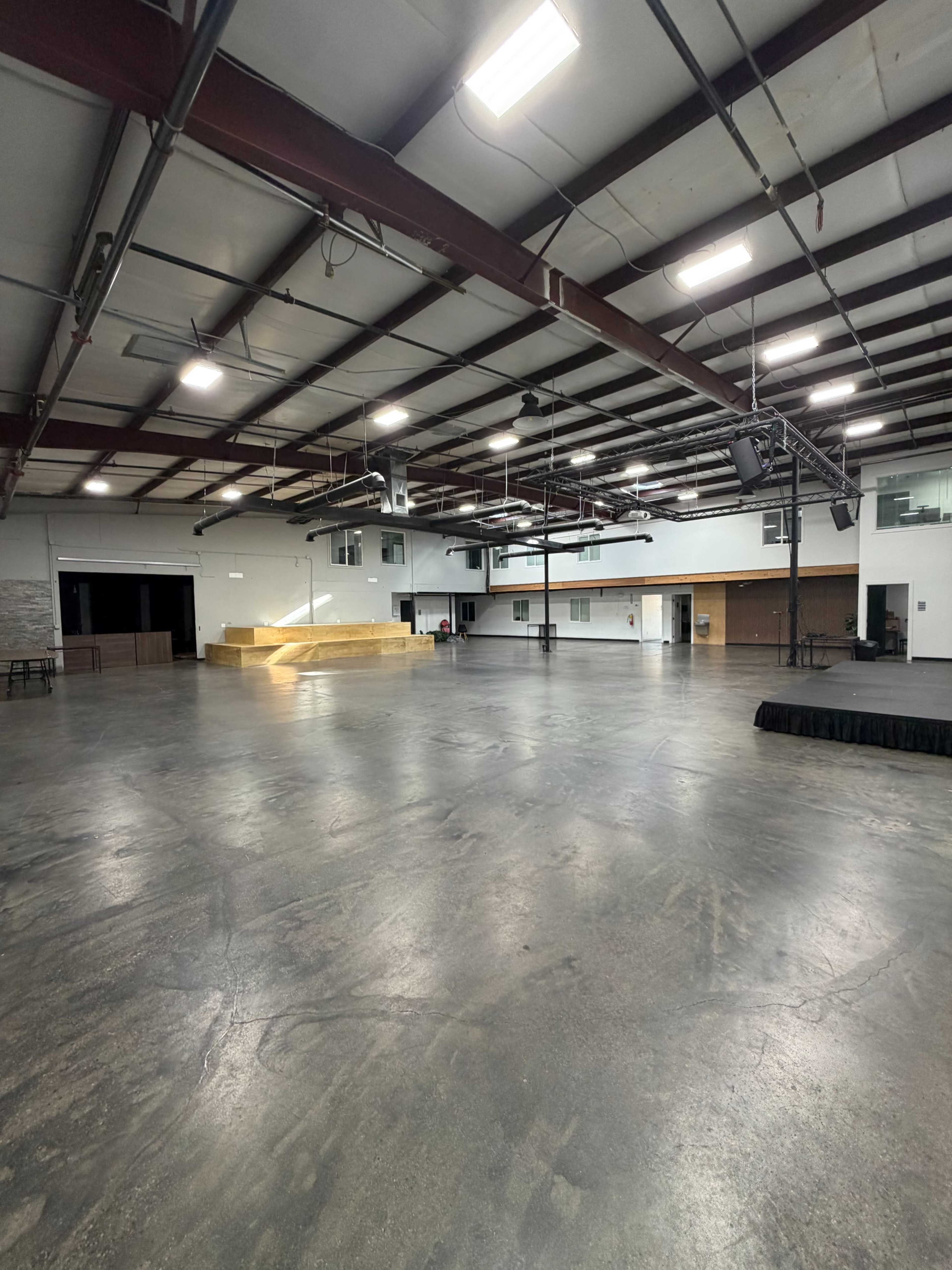 The image shows a spacious indoor venue with high ceilings, large open areas, and a wooden stage at one end.