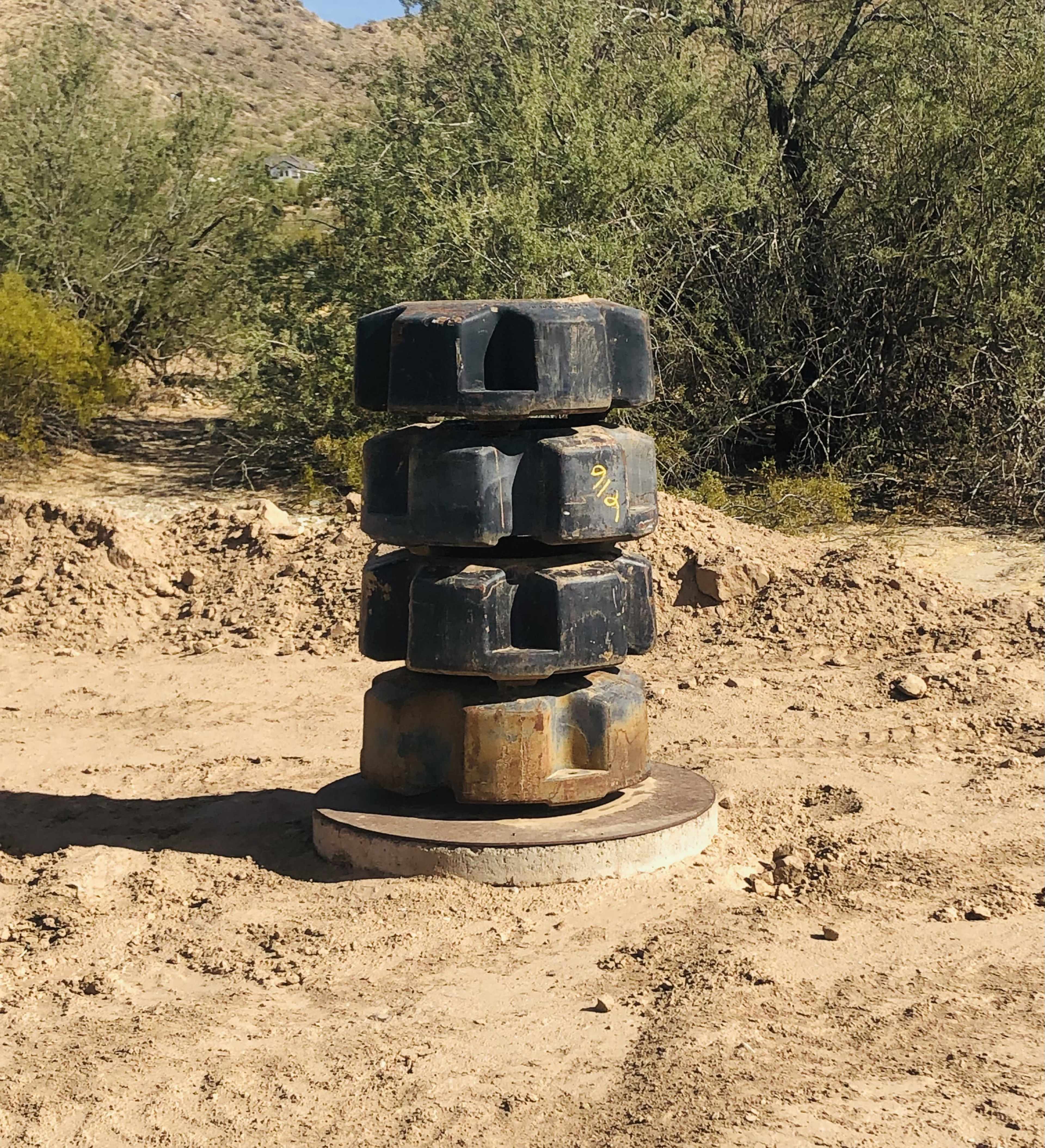 A stack of four large, worn rubber tires is positioned on a circular metal base in a desert landscape surrounded by sparse vegetation.