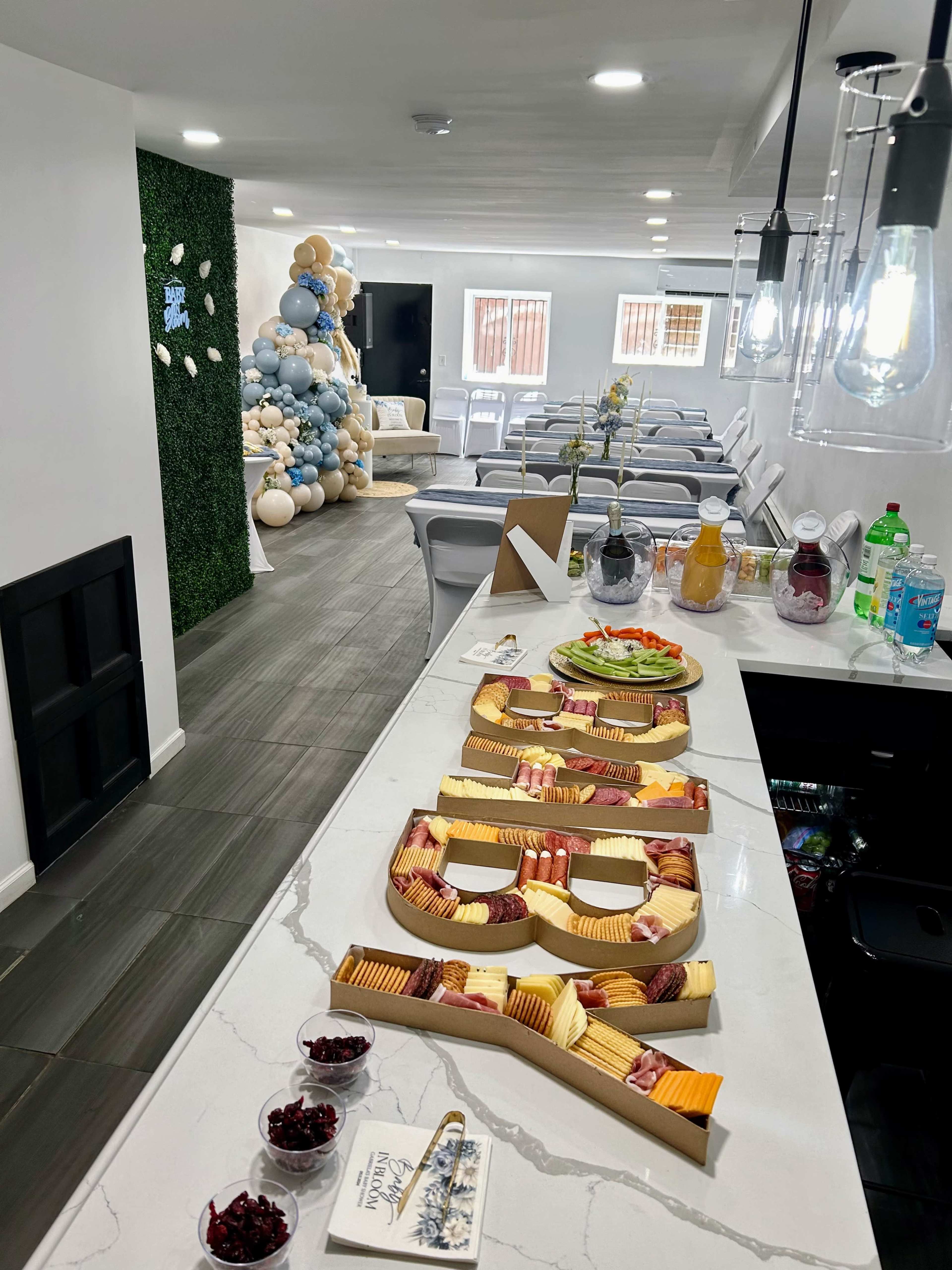 A long table displays a charcuterie board with various cheeses and meats, while a seating area and a balloon display are visible in the background.