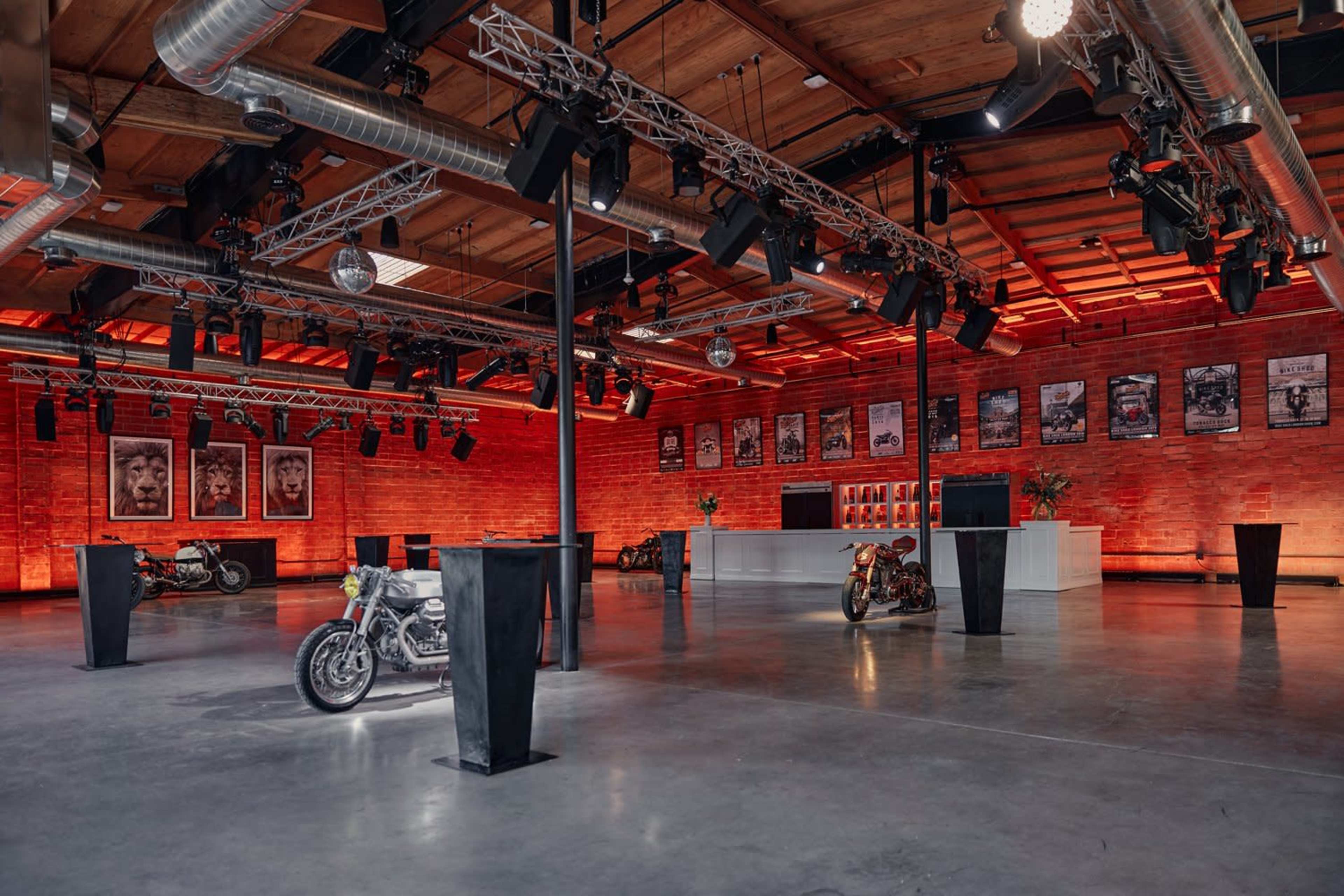 The spacious industrial venue features vintage motorcycles on display and walls adorned with posters, illuminated by red lighting.
