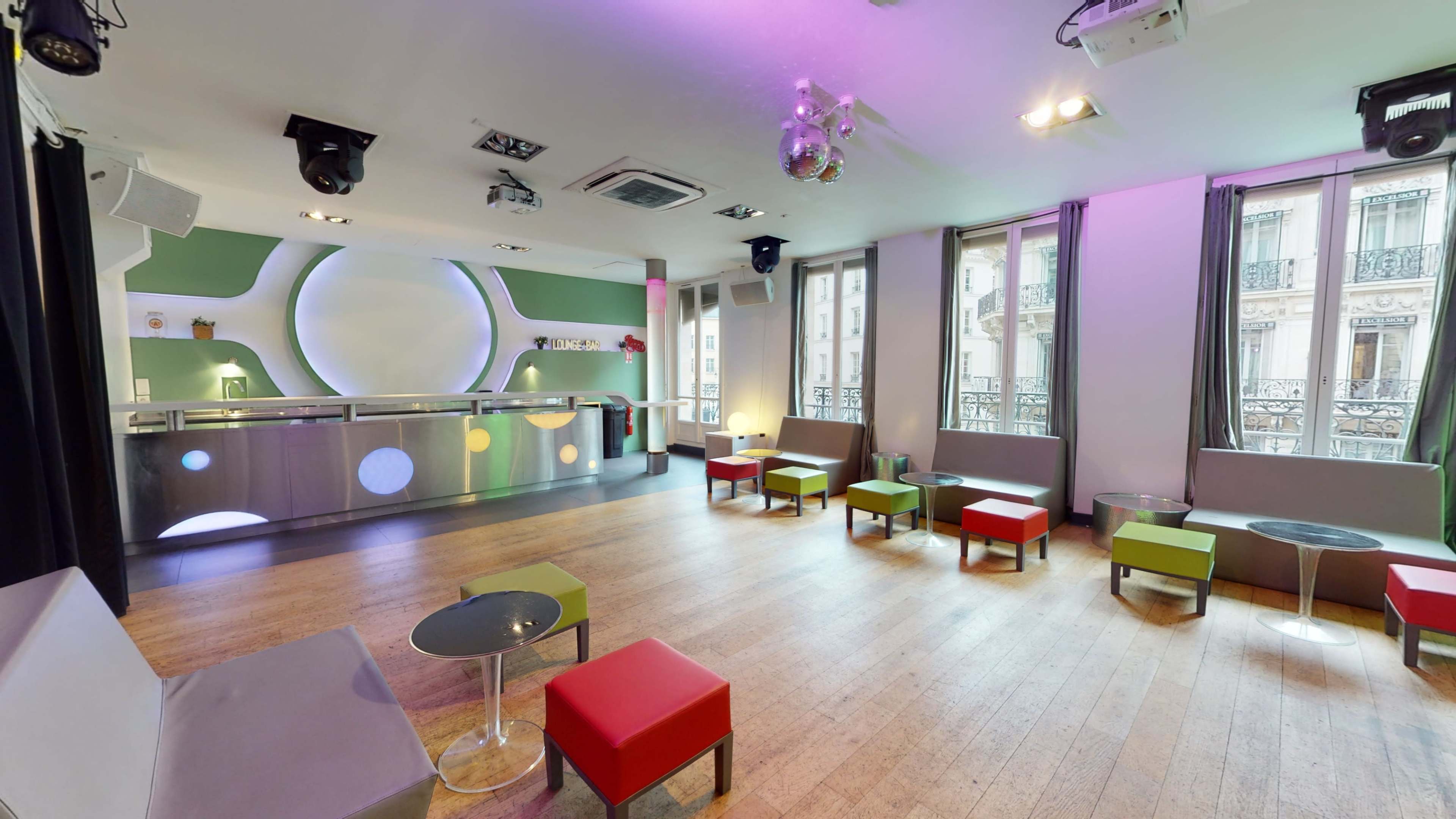 A modern lounge area with minimalist furniture, featuring a bar counter, colorful seating arrangements, and large windows allowing natural light to enter.