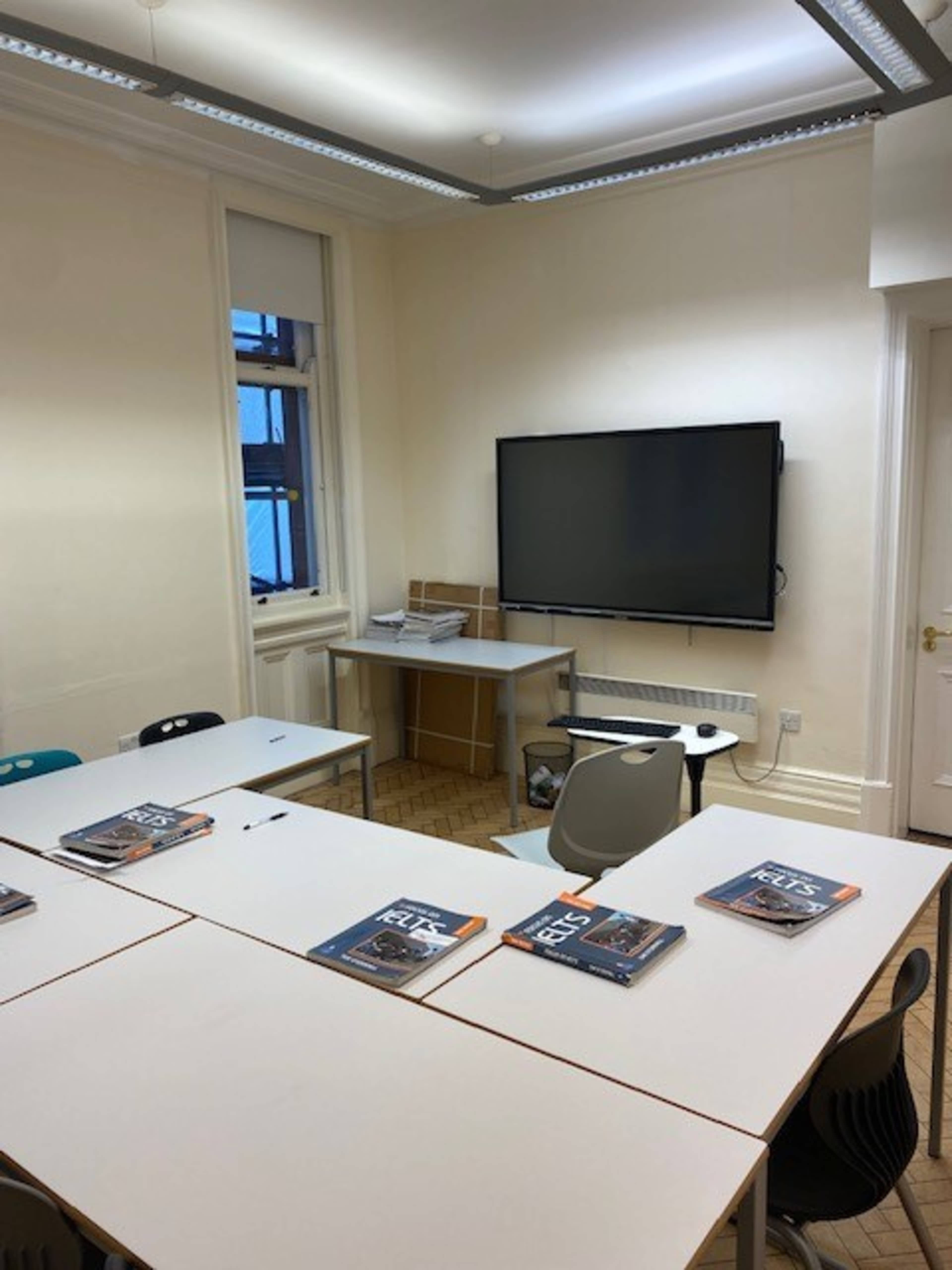 A classroom is arranged with several tables and chairs, along with a TV mounted on the wall and textbooks placed on the tables.
