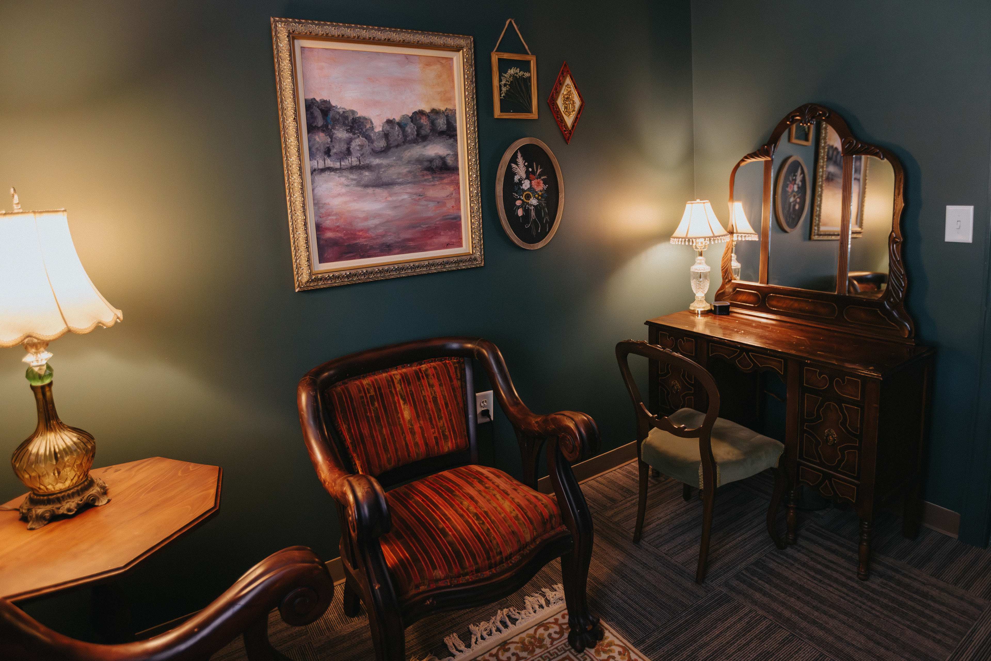 A small room with dark green walls, featuring a vintage chair, a wooden vanity with a mirror, and a decorative painting, illuminated by two lamps.