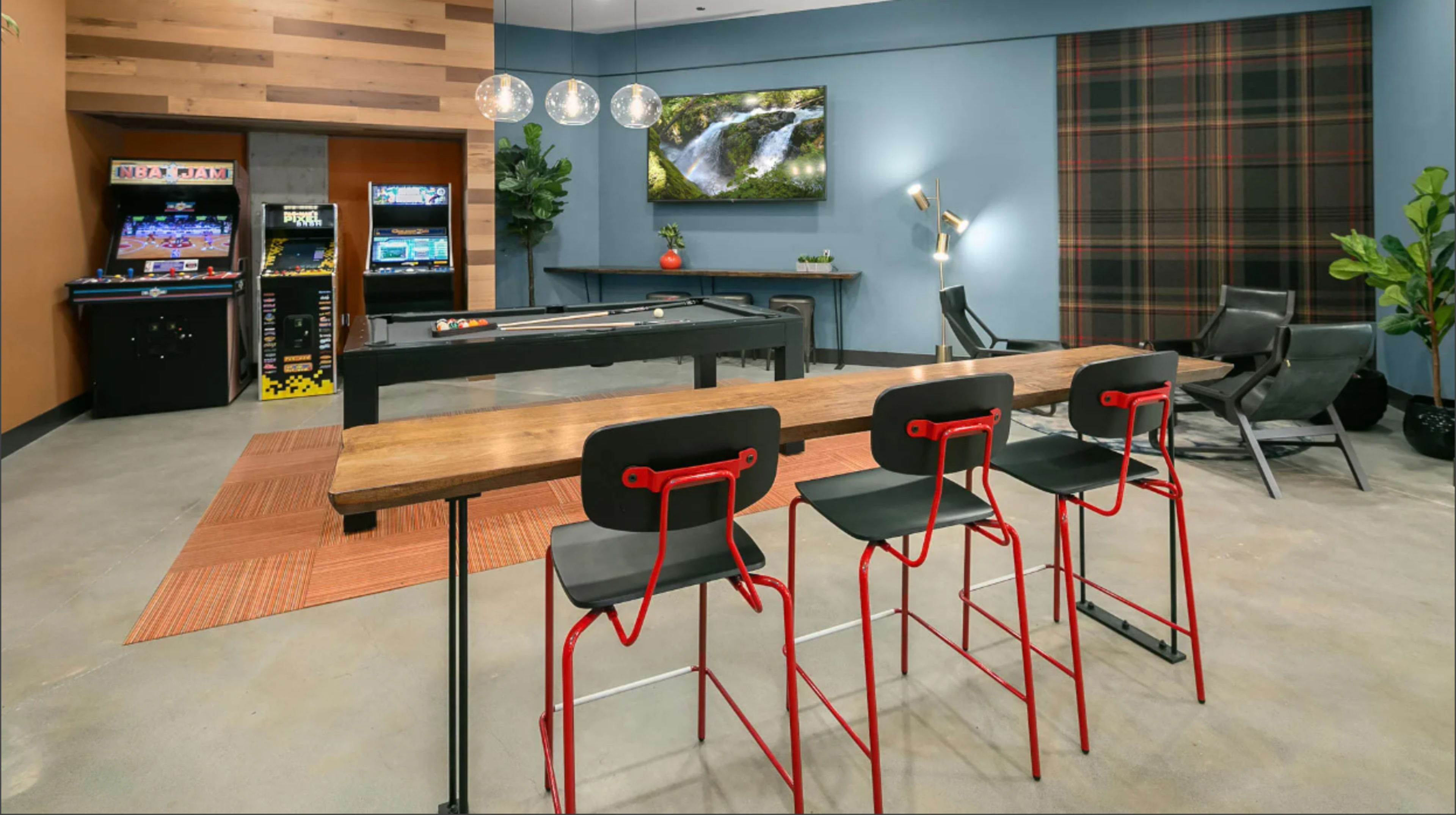The image shows a modern recreational room featuring a long table with red chairs, a large screen displaying a nature scene, and arcade games in the background.