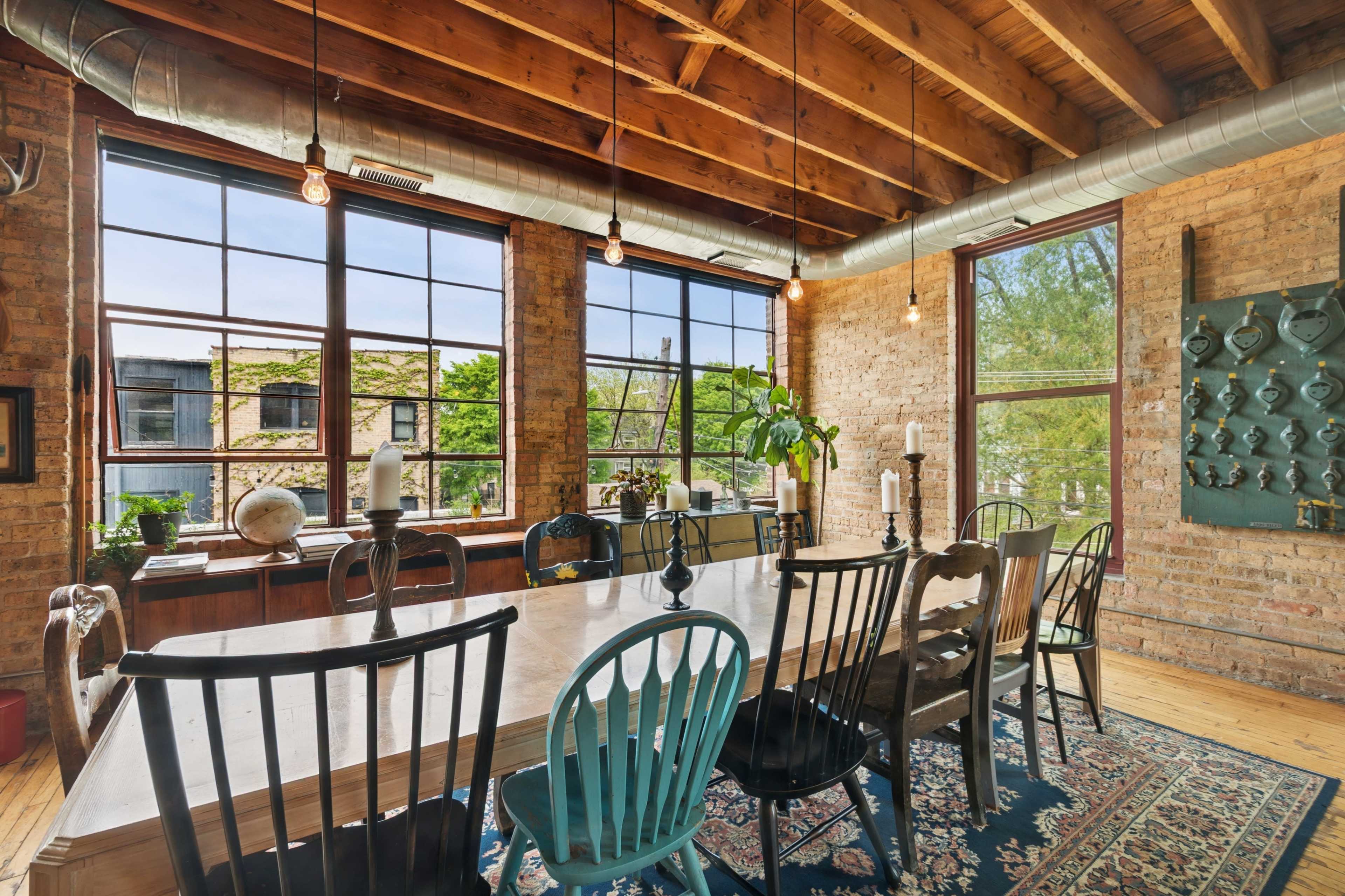 A spacious dining area features a long wooden table surrounded by mismatched chairs, with large industrial windows allowing natural light to fill the room.