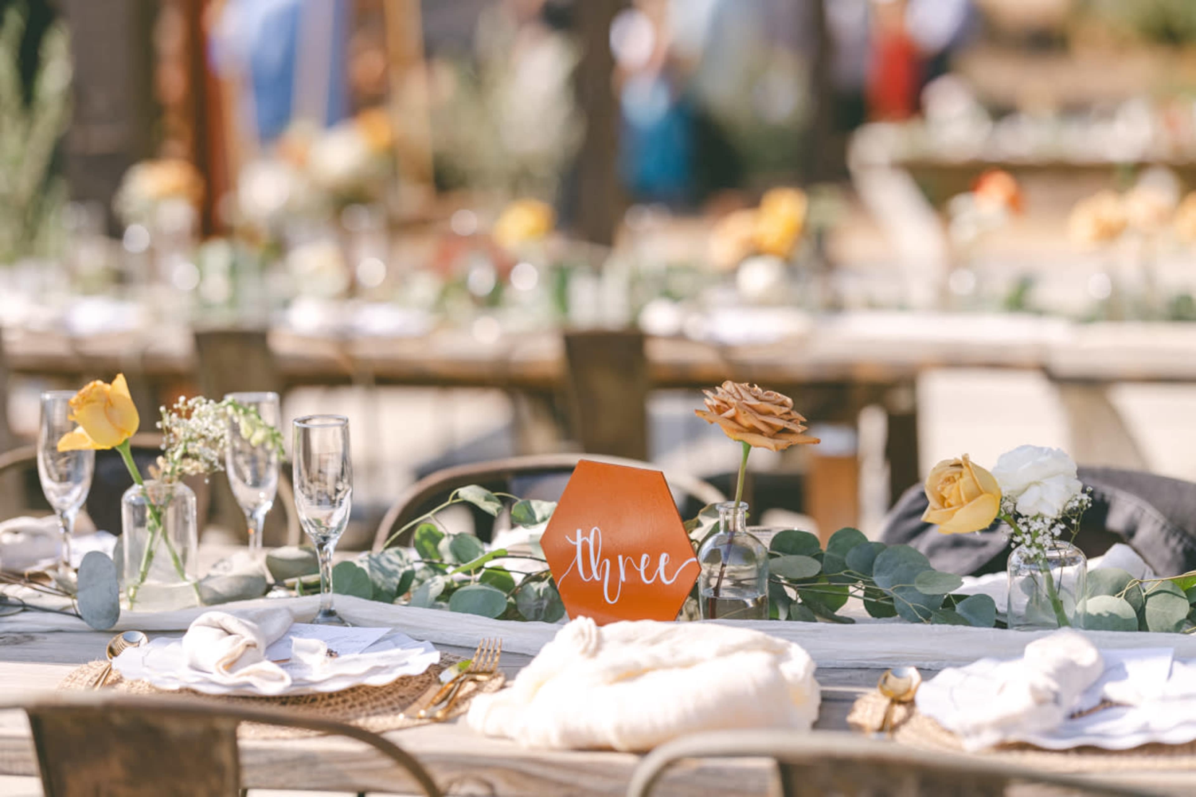 A rustic outdoor dining setup features long wooden tables adorned with floral centerpieces, napkins, and an orange table card labeled "Three."