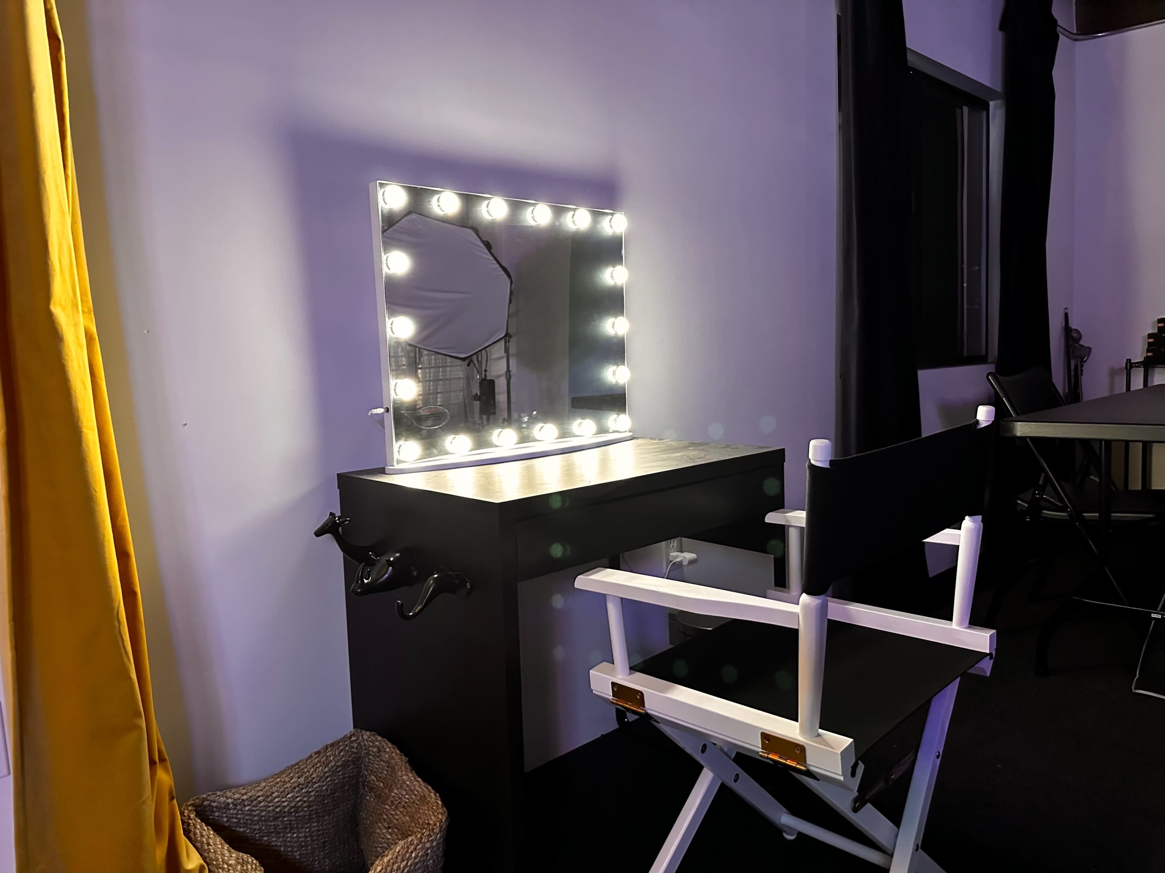 A makeup vanity with a lighted mirror next to a director's chair in a room with dark walls.