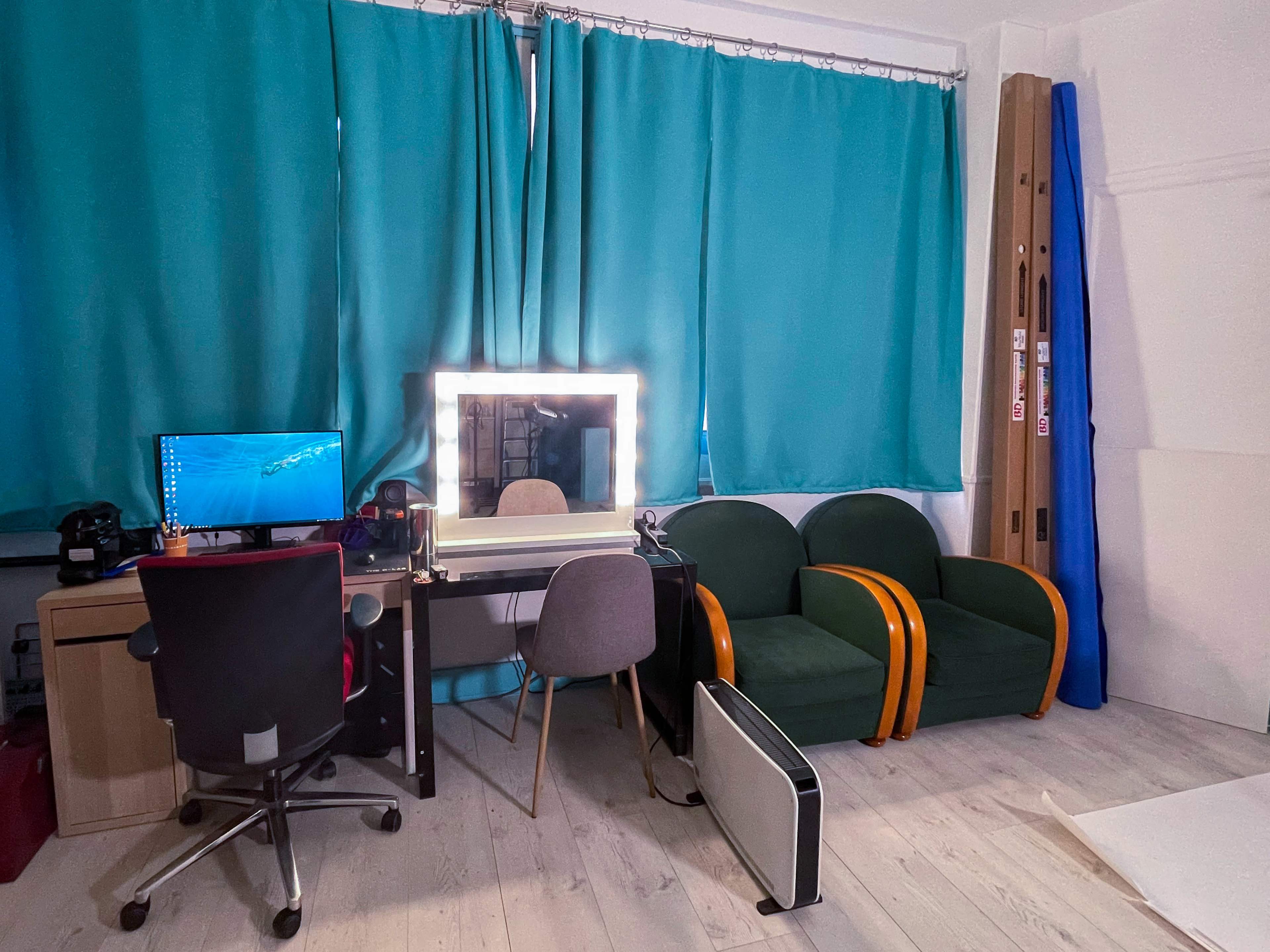 A workspace featuring a desk with a computer, a mirror with lights, two green chairs, and a heater, against a backdrop of turquoise curtains.