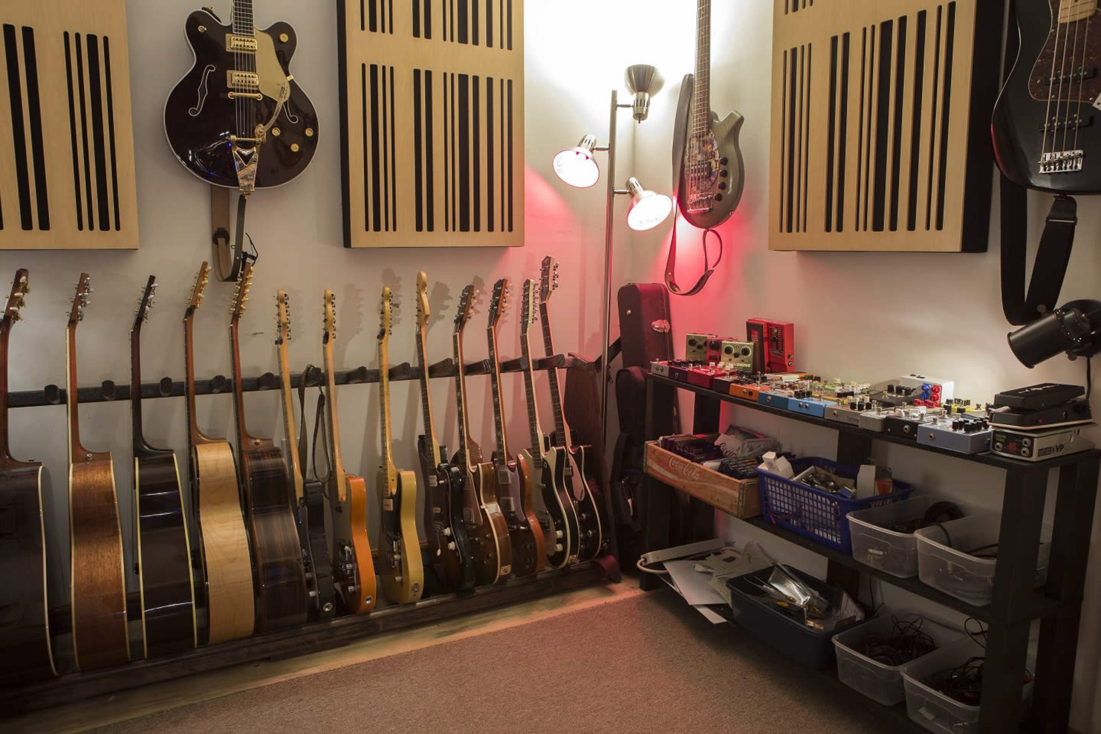 The image shows a room arranged for music production, featuring a rack of various guitars on one side and a table with an assortment of audio effects pedals and equipment on the other.