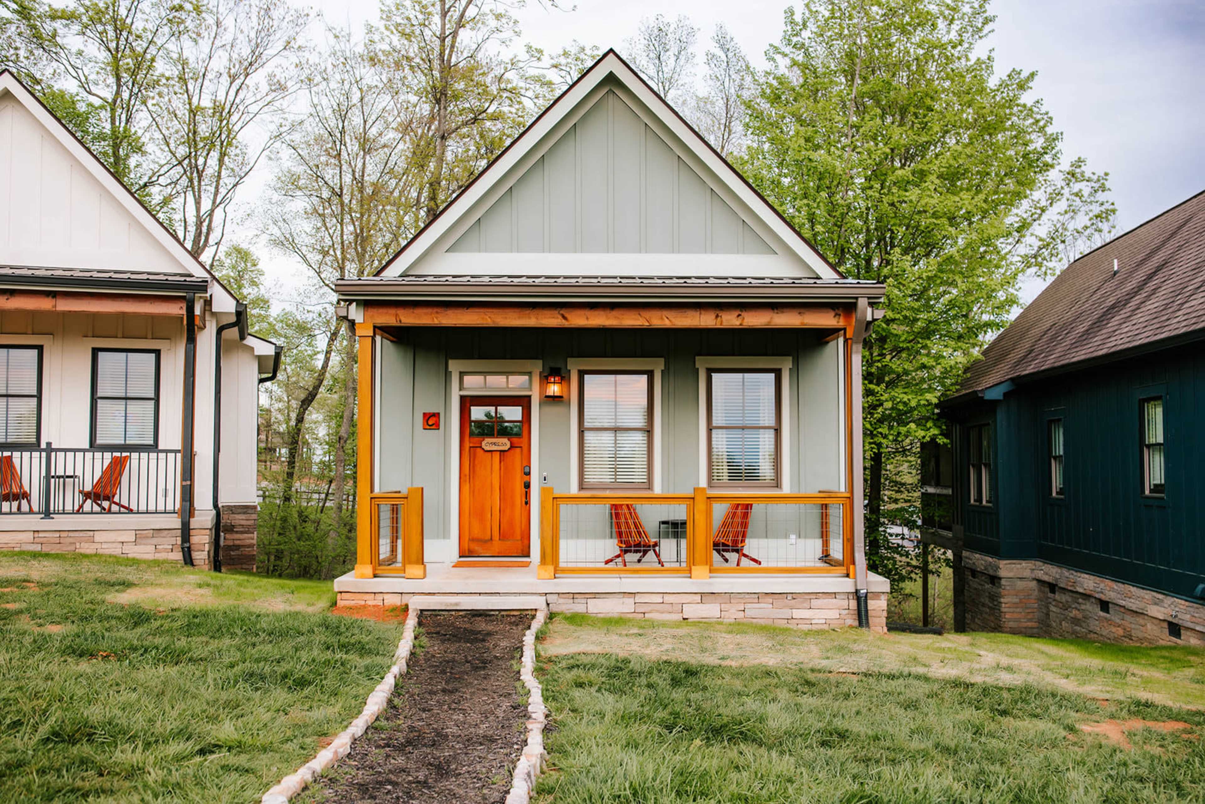 A small, light green house with a front porch and two chairs sits among trees and grassy landscaping.